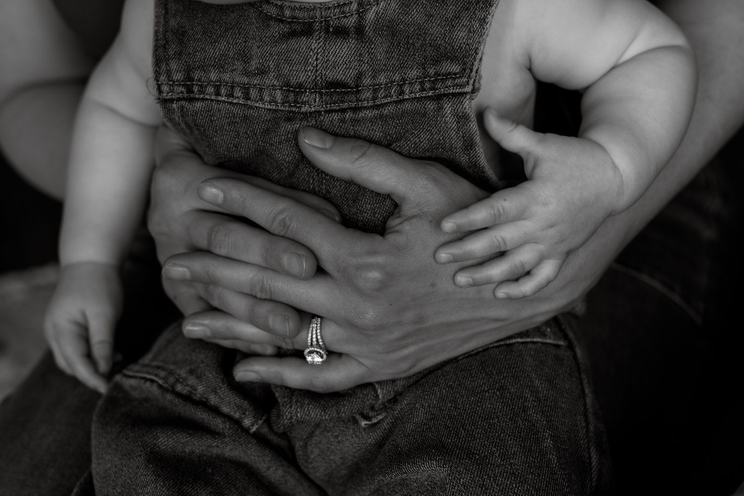 Black and white photo of adult hands cradling a baby sitting on their lap, wearing denim overalls.