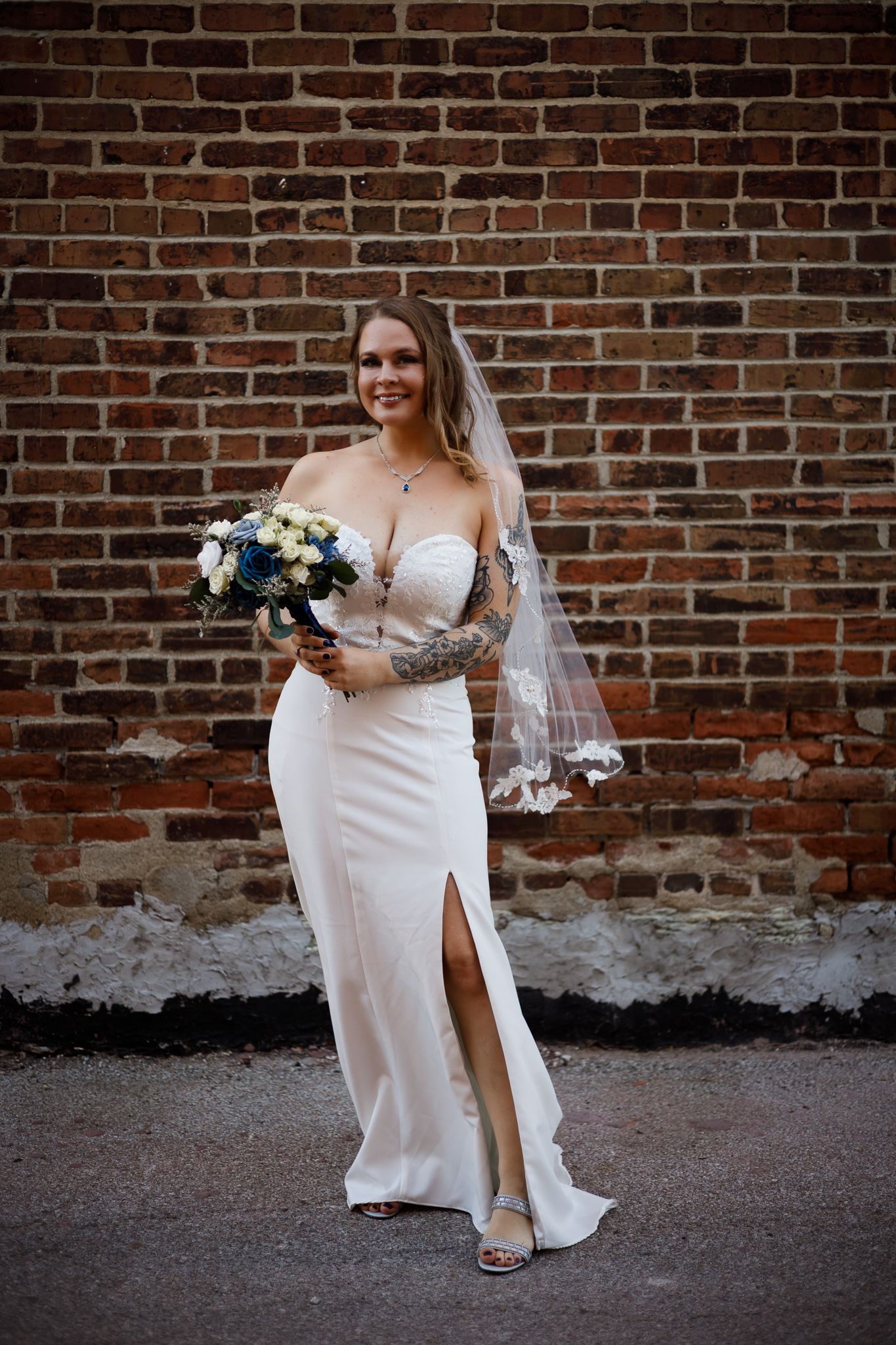 A bride in a white strapless gown poses against a rustic brick wall while holding a blue and white floral bouquet.