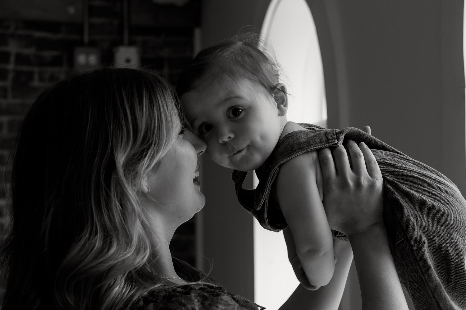 Black and white photo of a woman lovingly holding a baby face-to-face in a warmly lit room.
