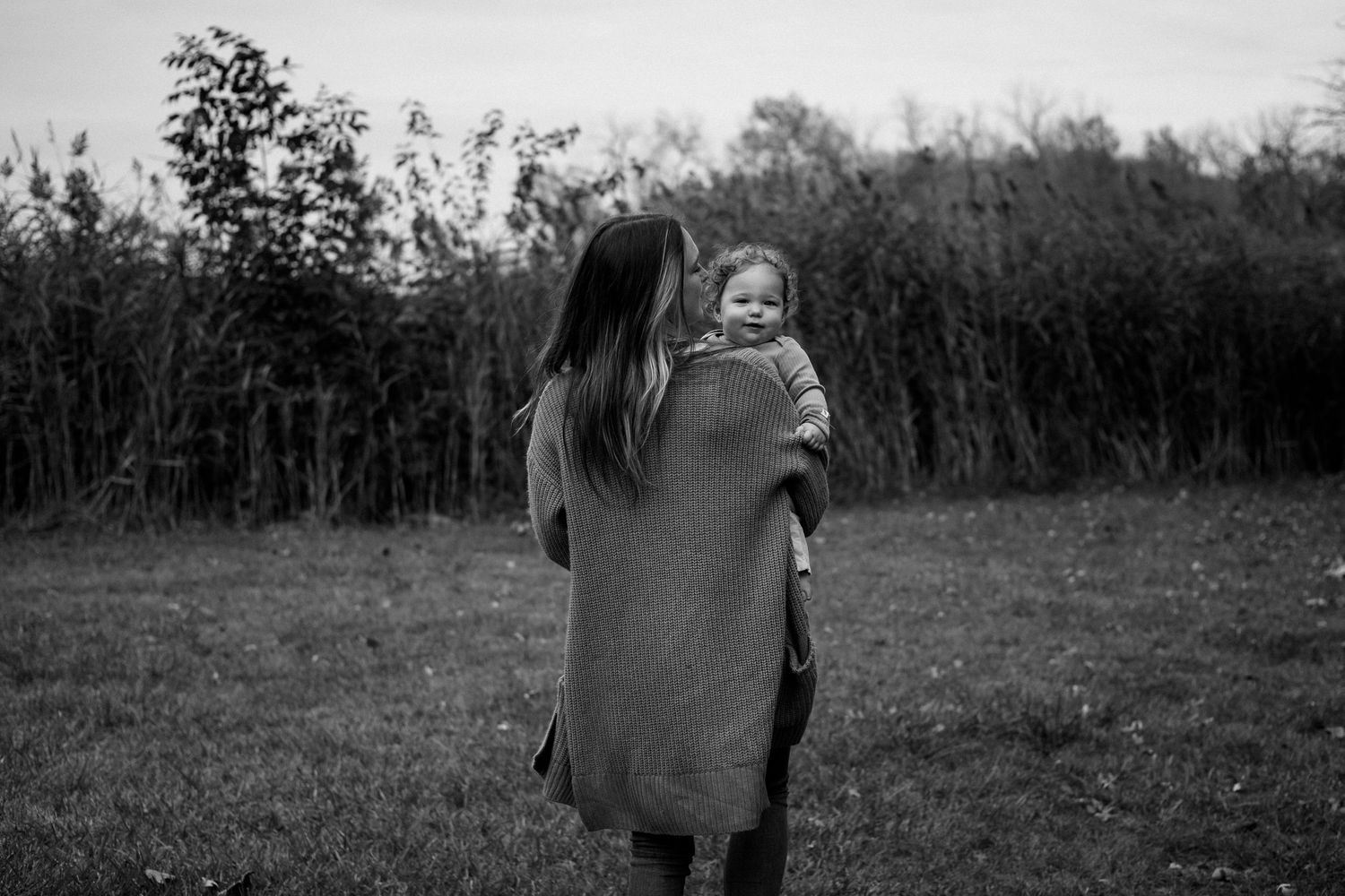 Black and white photo of someone in a sweater holding a small child outdoors in a natural setting.