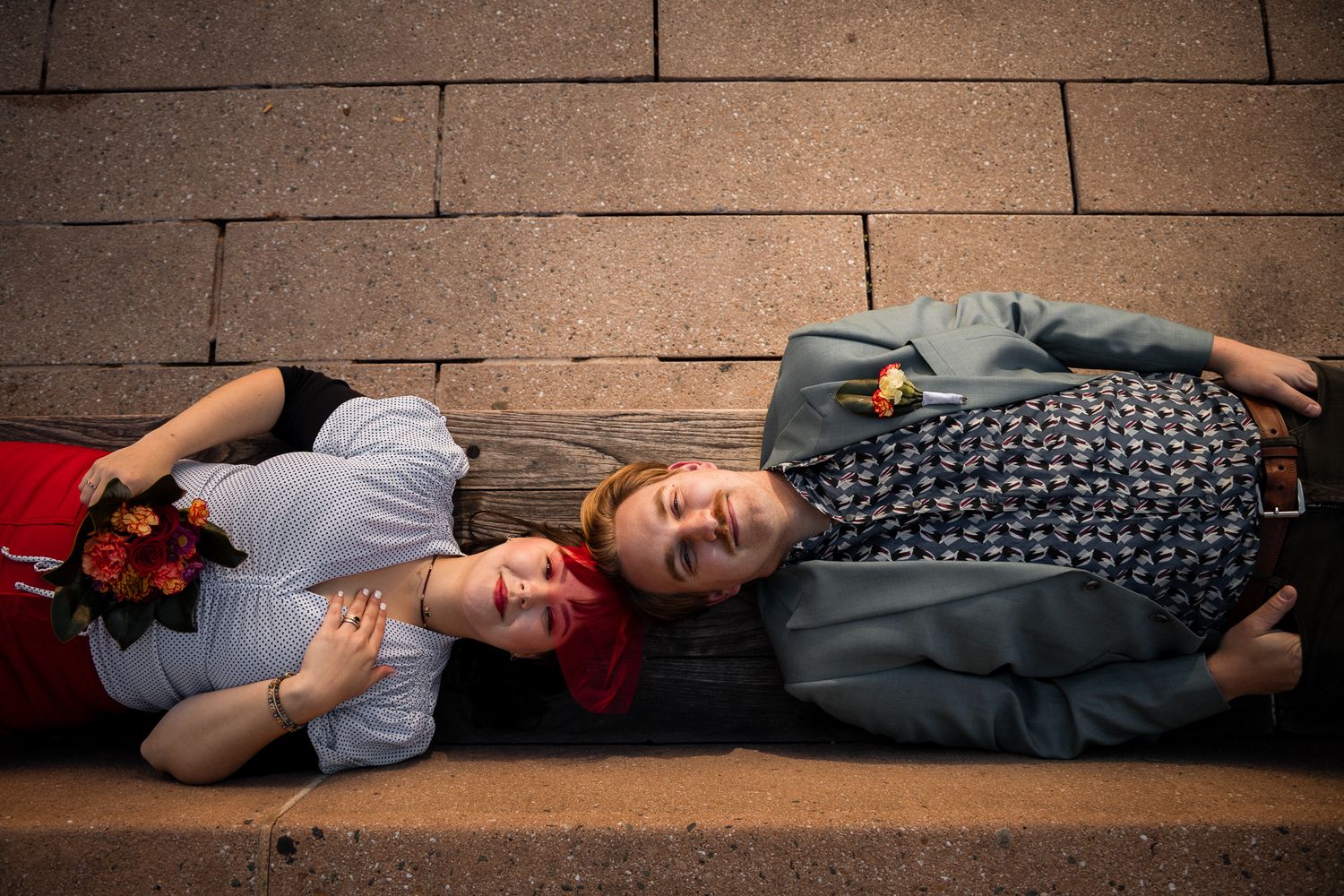 Couple relaxing and laughing while lying on a wooden bench against stone wall backdrop.