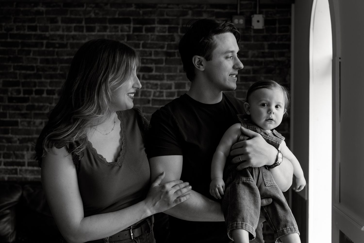Black and white photo of a young family, parents smiling with their baby boy near a brick wall window.