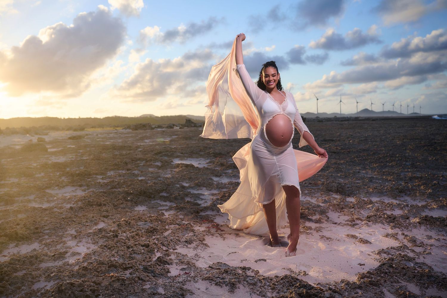 Pregnant woman in flowing white dress celebrating outdoors at sunset.