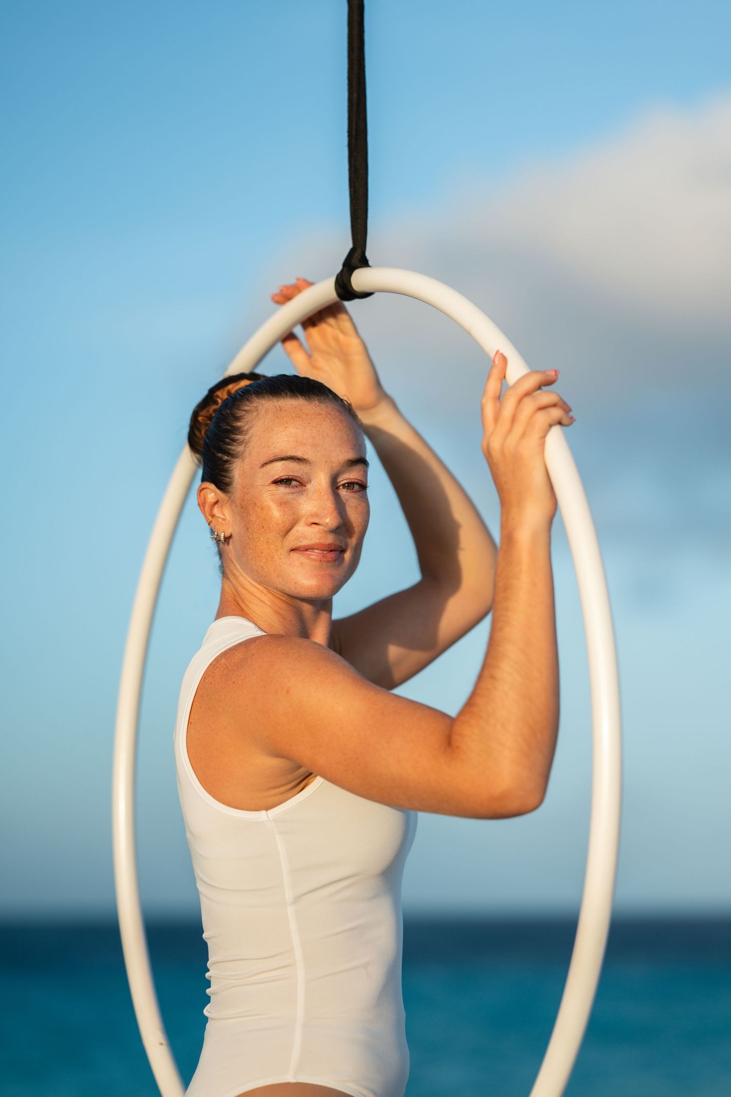 Woman smiling while holding aerial hoop on beach at golden hour.