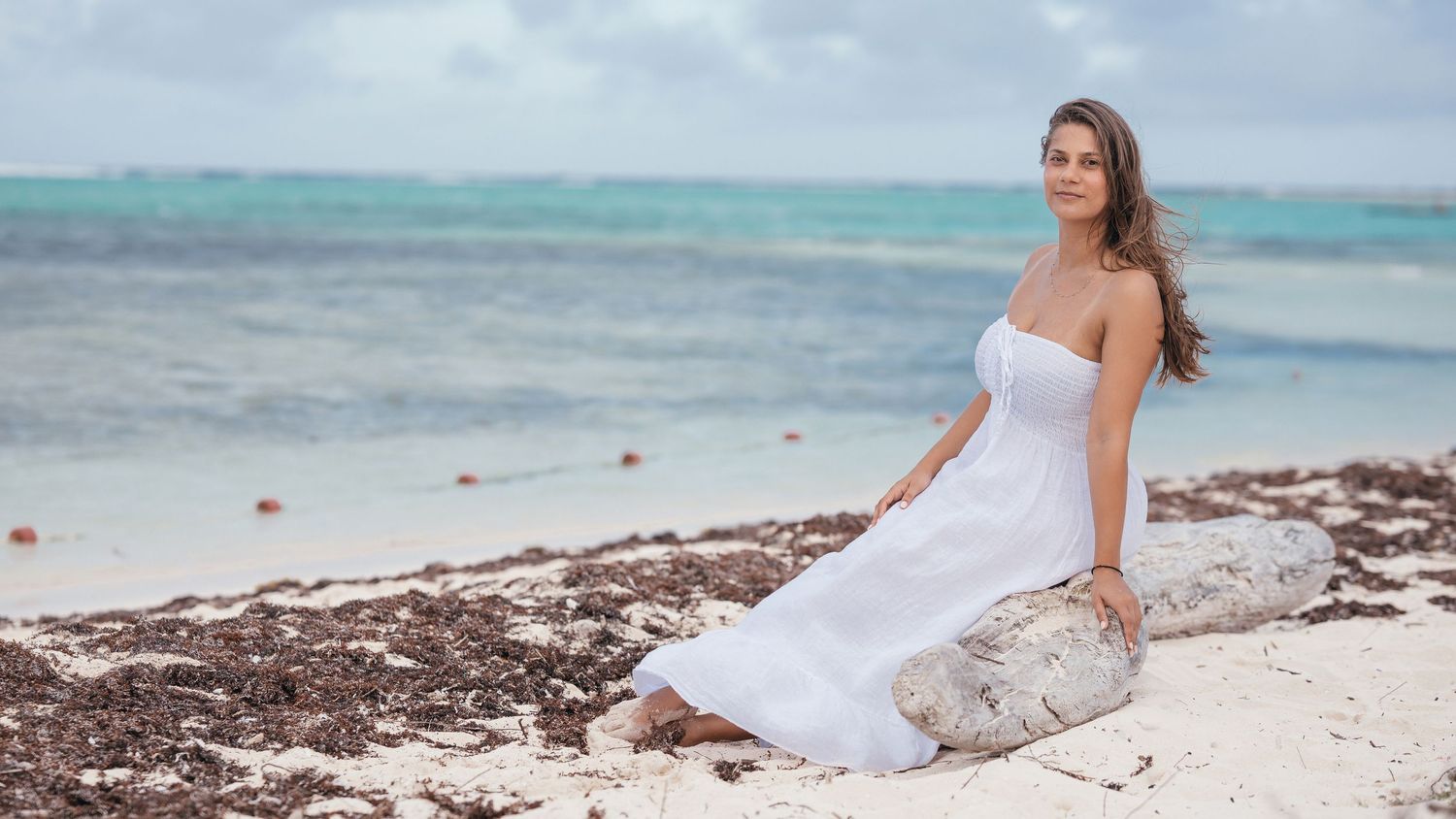 Woman in white strapless dress sitting on driftwood on tropical beach with turquoise sea.