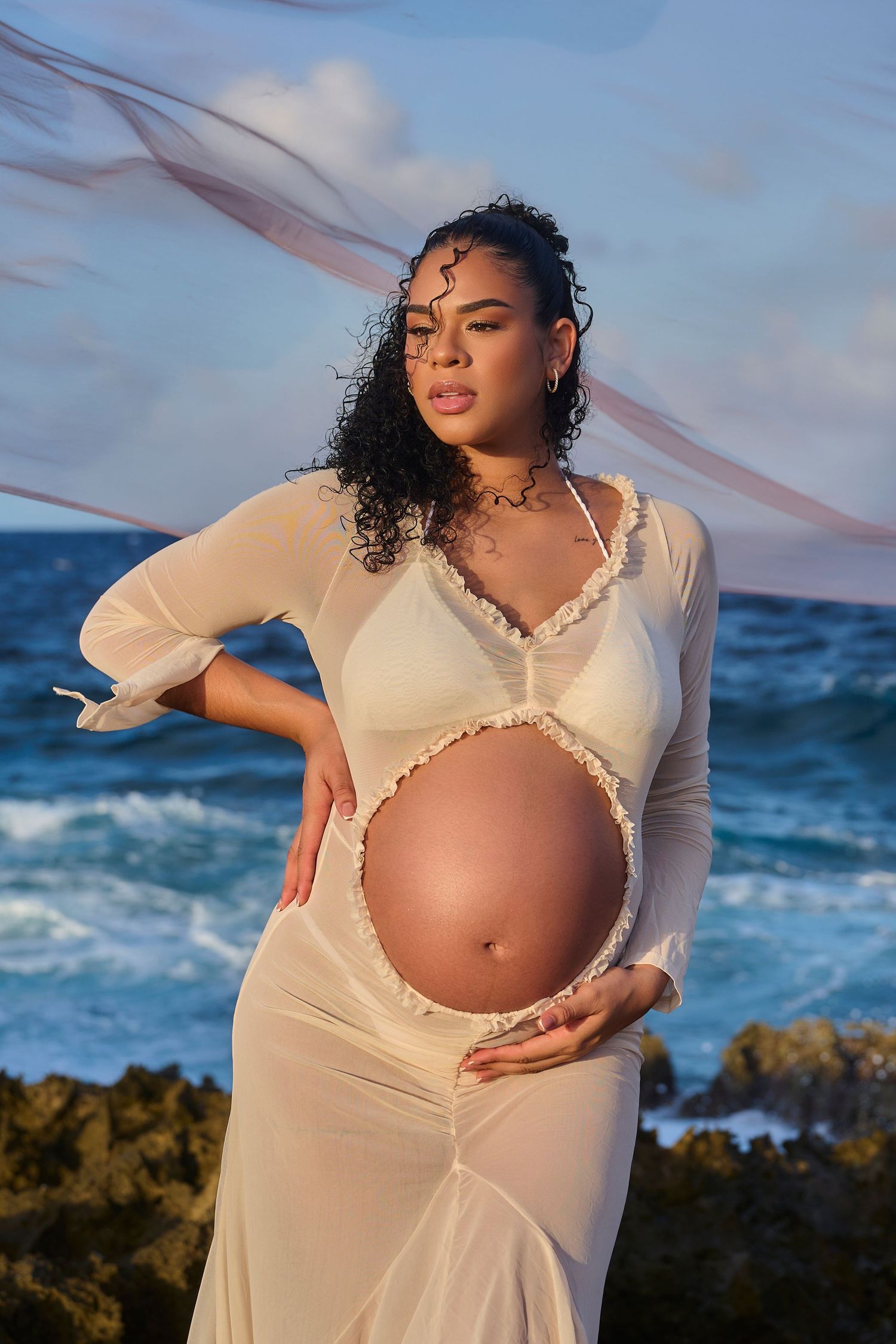 Beautiful pregnant woman in white lace maternity dress stands on ocean rocks at golden hour.