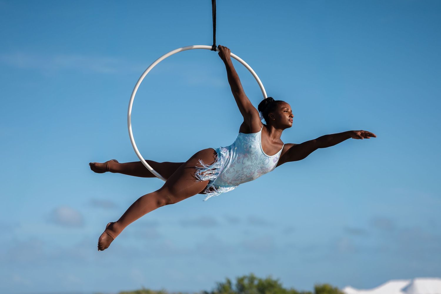 Female aerialist gracefully posing on aerial hoop against blue sky.