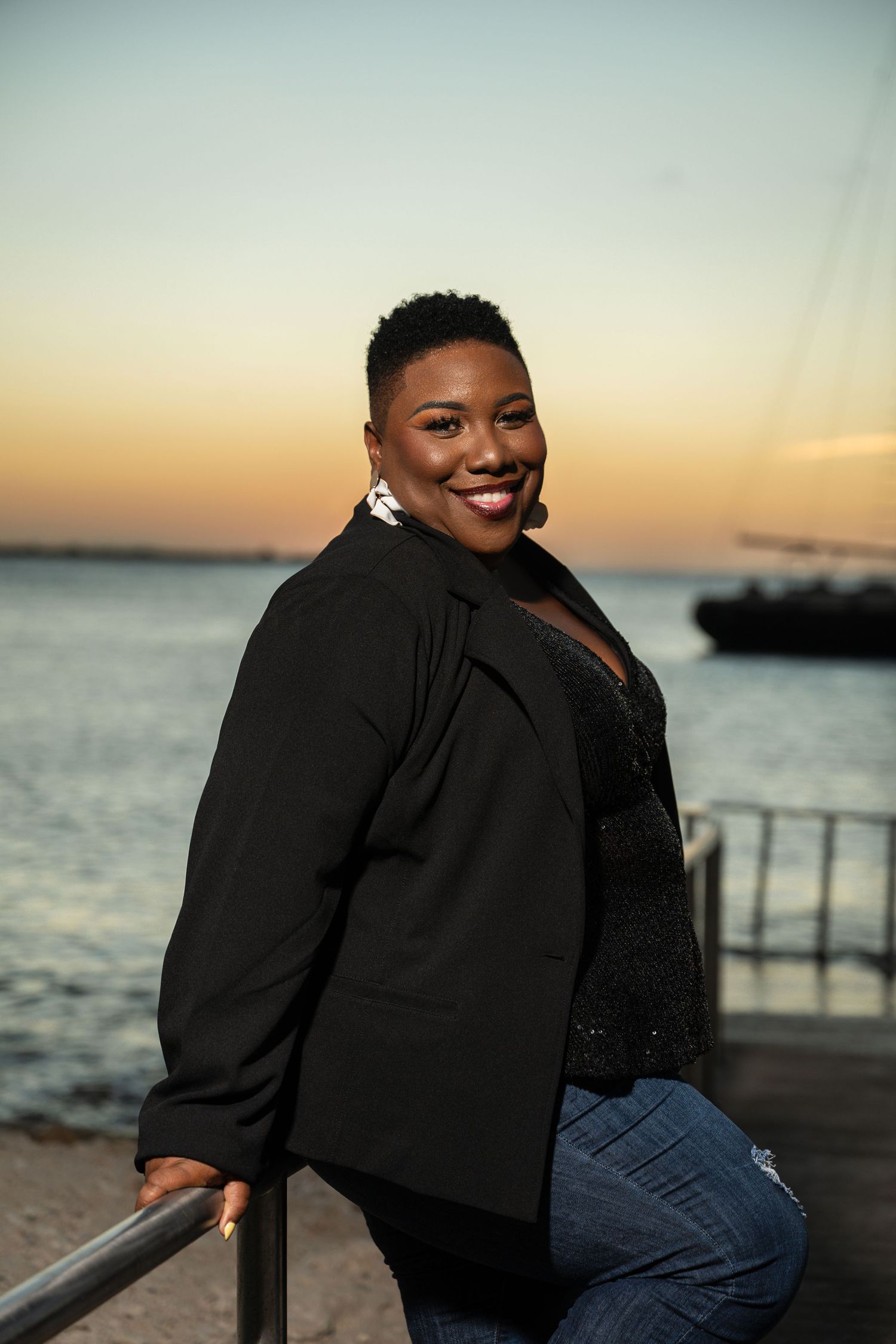 Confident Black woman in black blazer and jeans poses at waterfront railing during sunset.