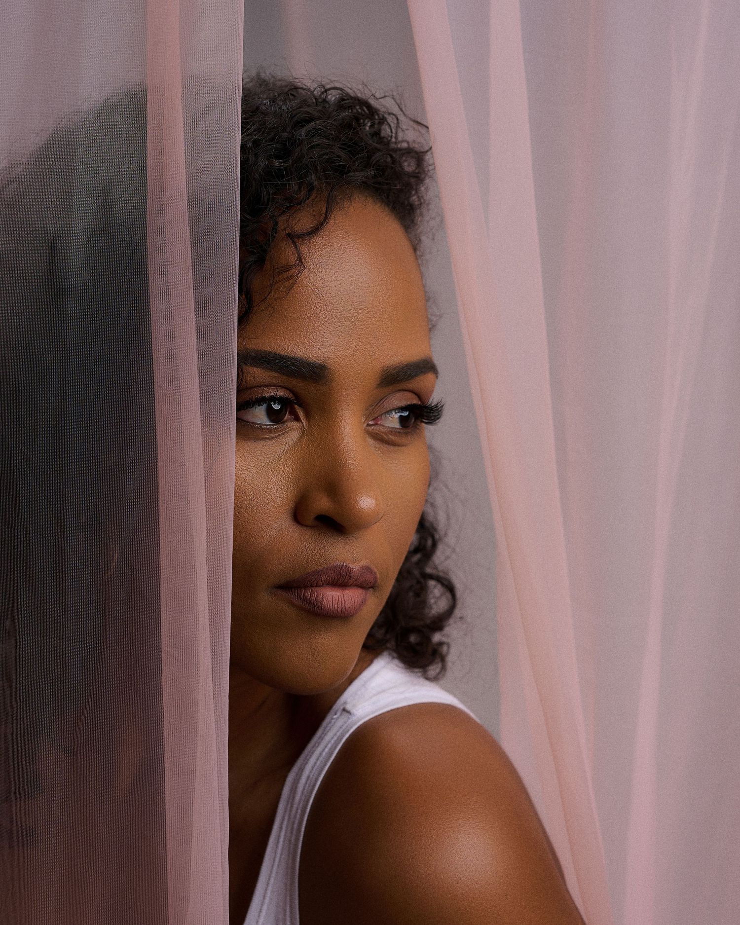 Close-up portrait of a woman with curly hair peeking through sheer pink curtains with a soft gaze.