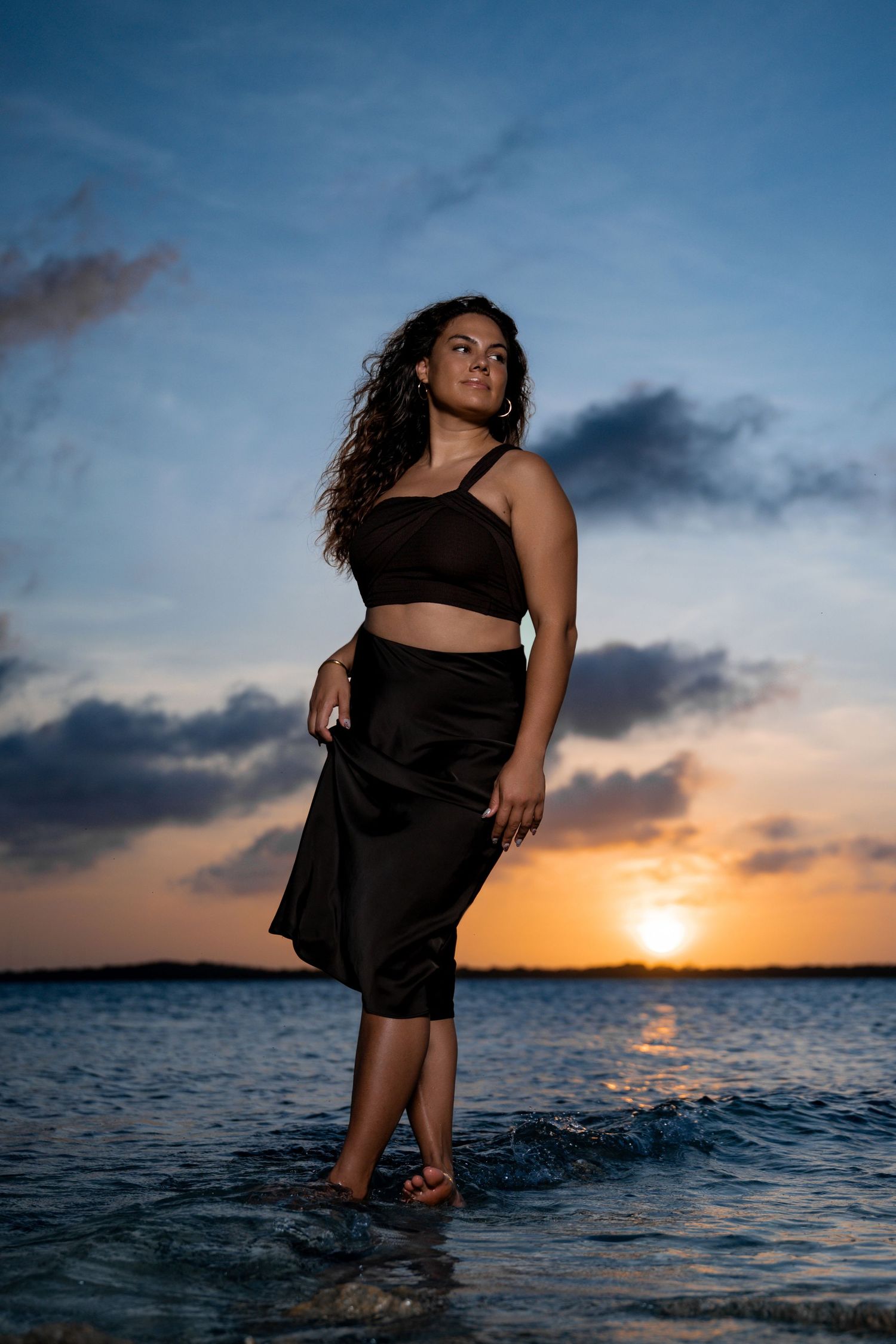 Woman in black crop top and skirt standing in ocean water at sunset with colorful sky.