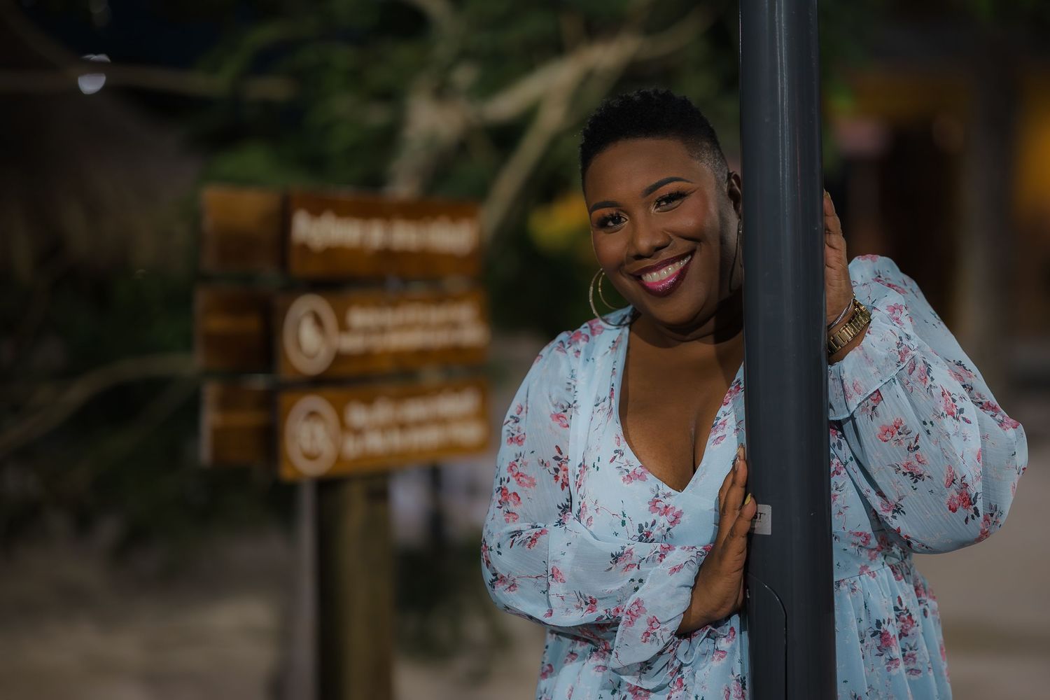 Smiling Black woman in floral dress leans against outdoor signpost at night.