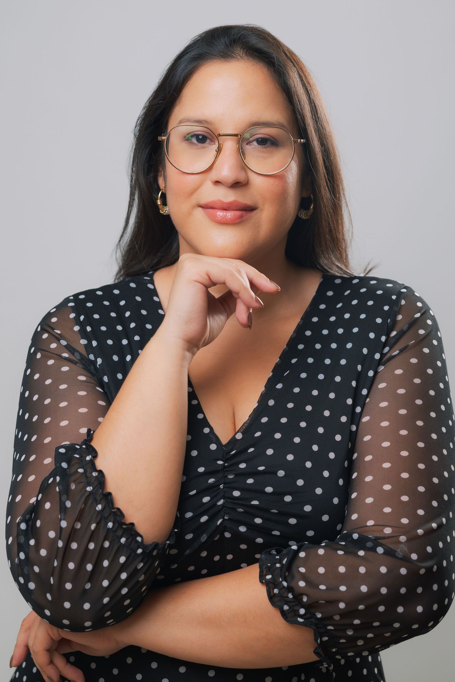 Woman with glasses in black polka dot dress poses thoughtfully with hand on chin in professional portrait.