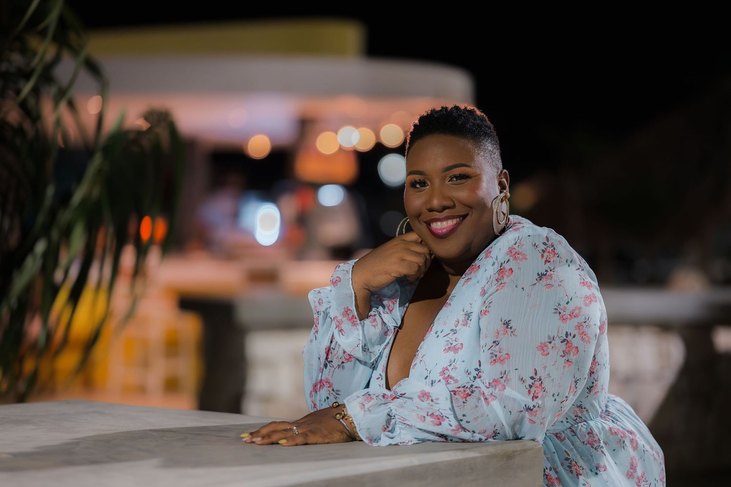 Happy Black woman in floral wrap dress smiles while leaning on white railing at night.
