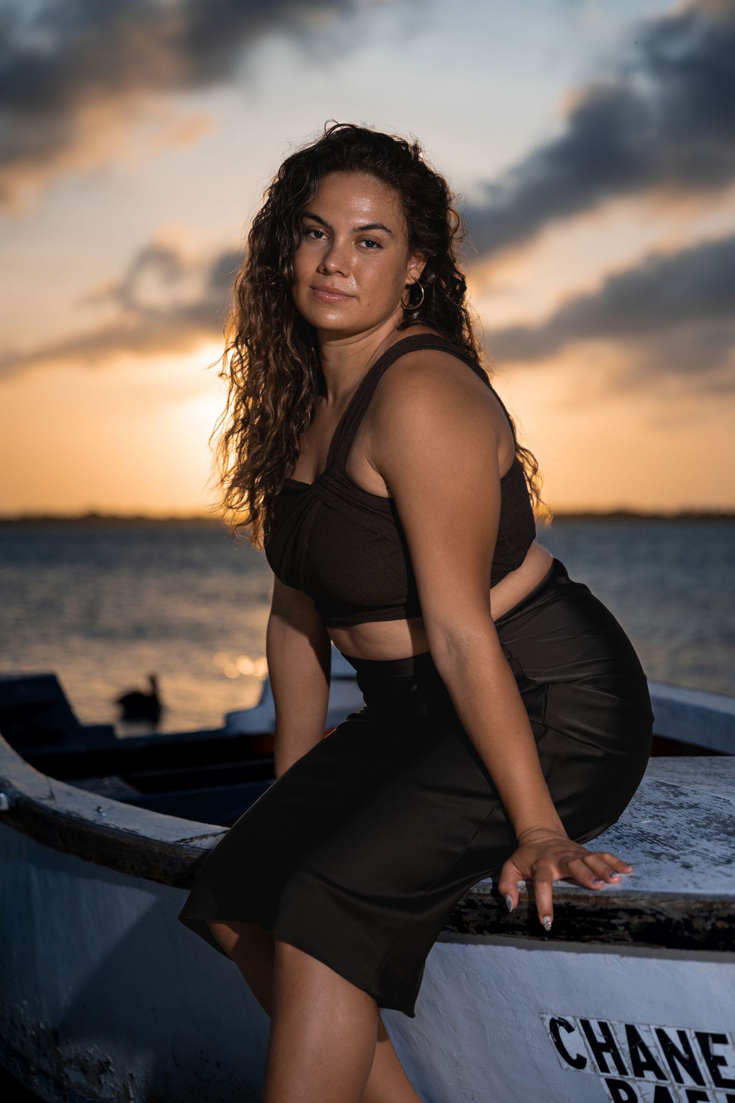 Woman in black two-piece outfit posing on white boat at golden sunset by the sea.