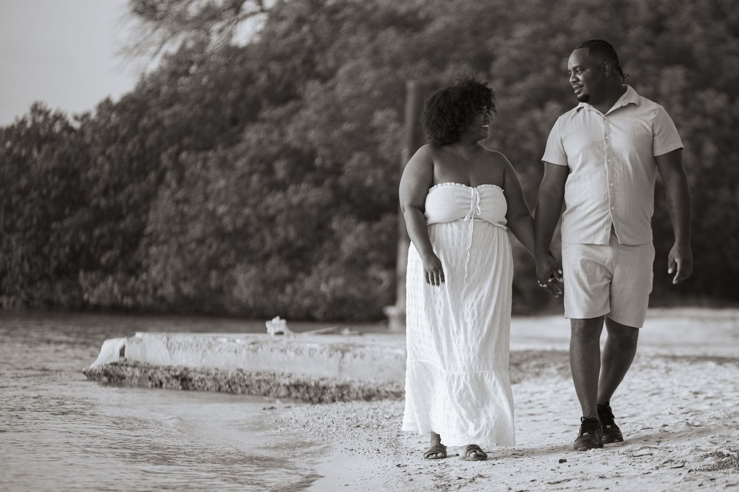 Black and white photo of couple holding hands walking on tropical beach near water.