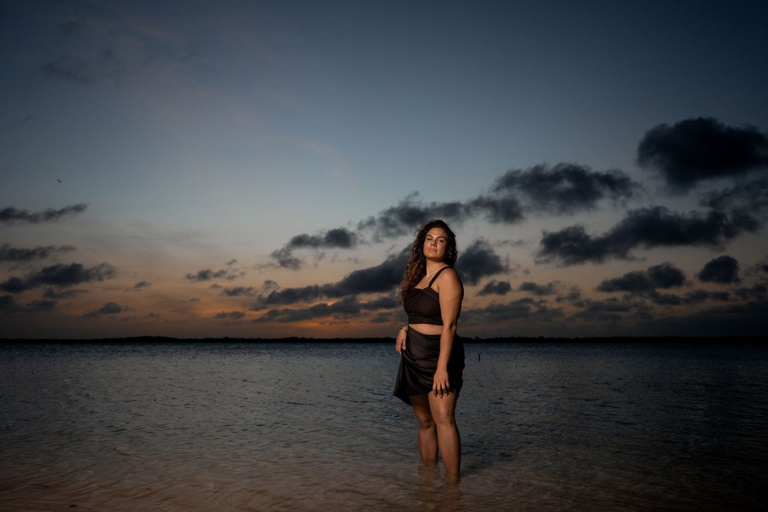 Woman in black outfit standing in shallow ocean water during dramatic sunset with cloudy sky.