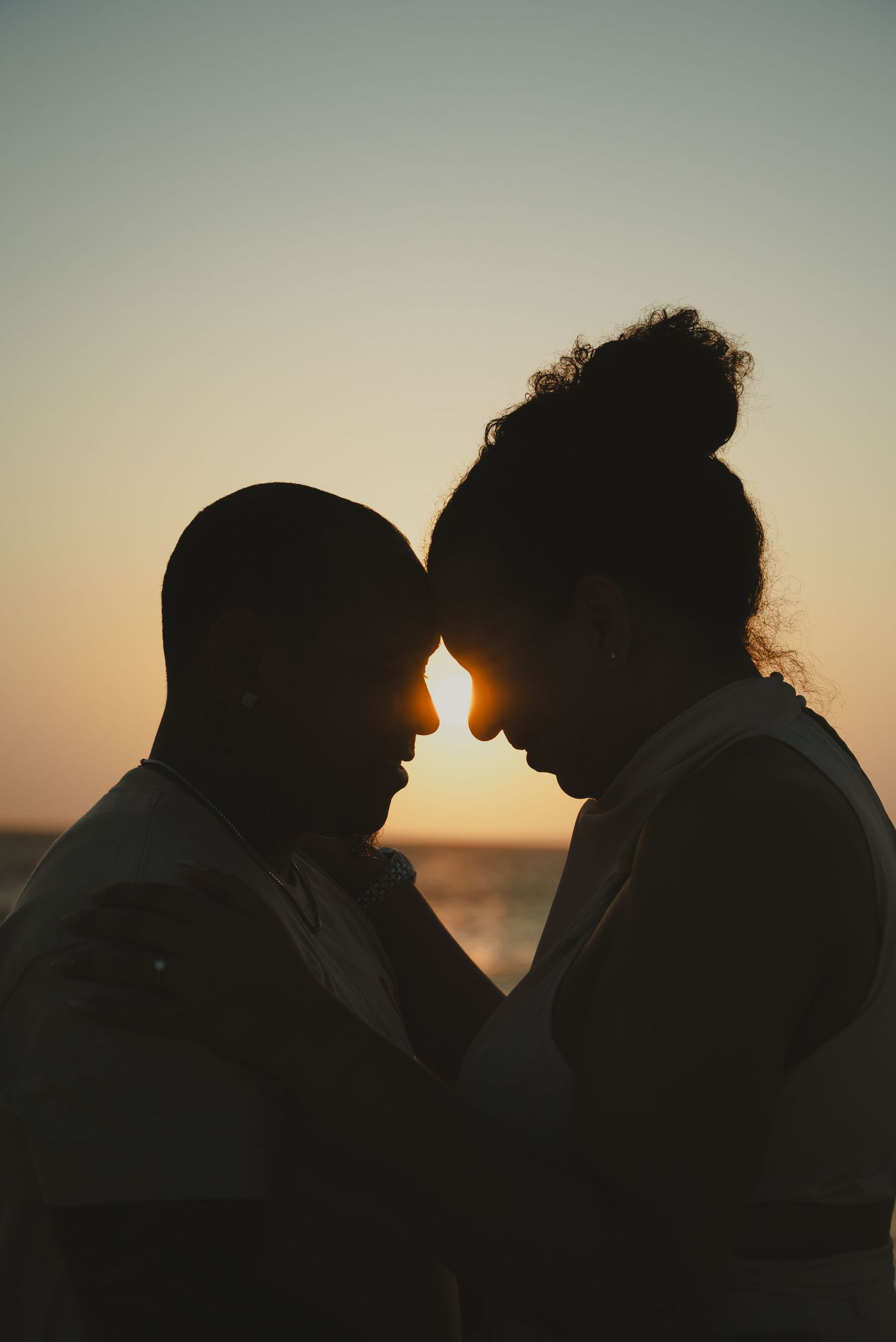 Romantic silhouette of couple touching foreheads against golden sunset sky near ocean.