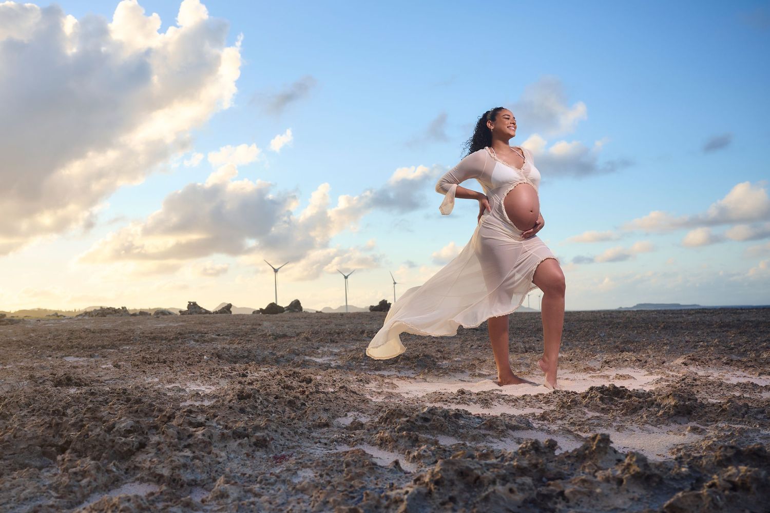 Pregnant woman in white dress posing on rocky beach at sunset.