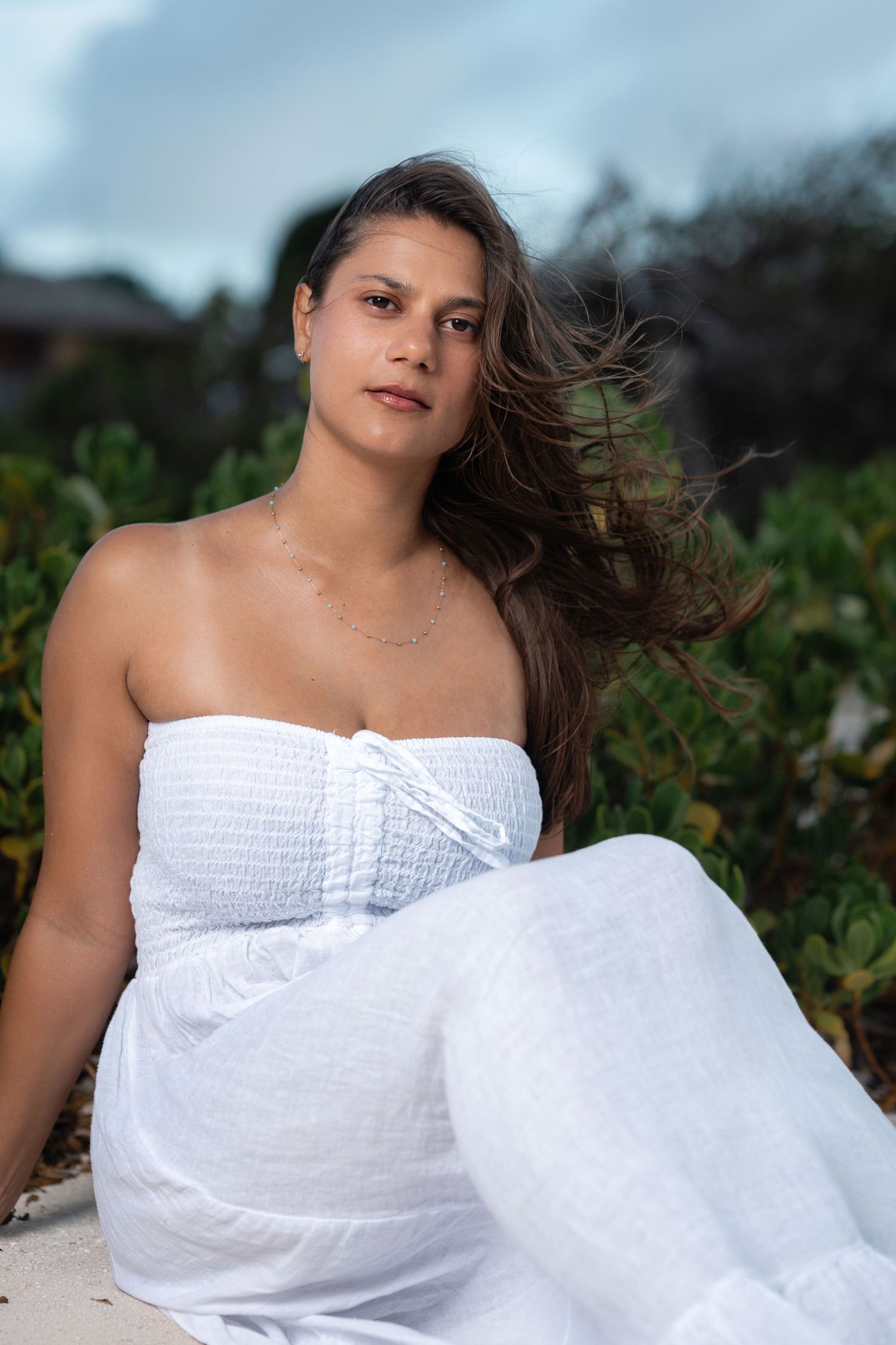 Young woman in white strapless dress sitting outdoors with hair blowing in wind.
