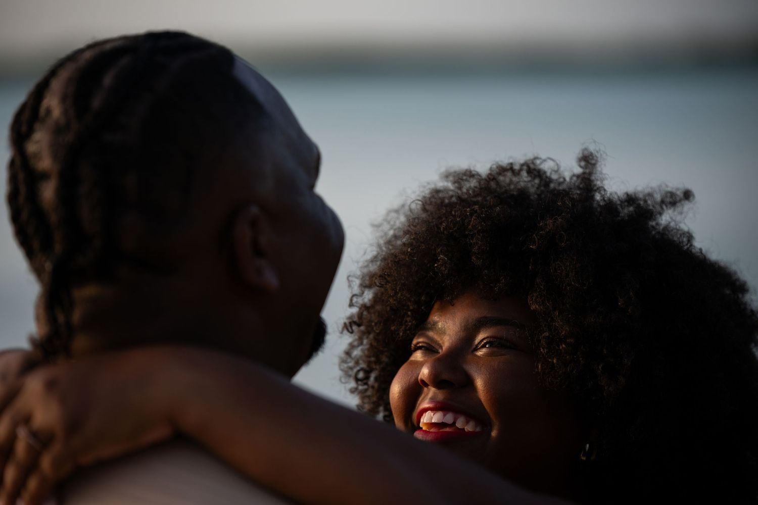 Joyful woman with curly hair laughing while embracing partner near sparkling ocean water.