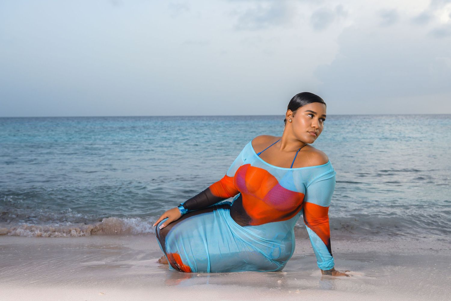 Woman in colorful blue and orange bodysuit poses on sandy beach by ocean.