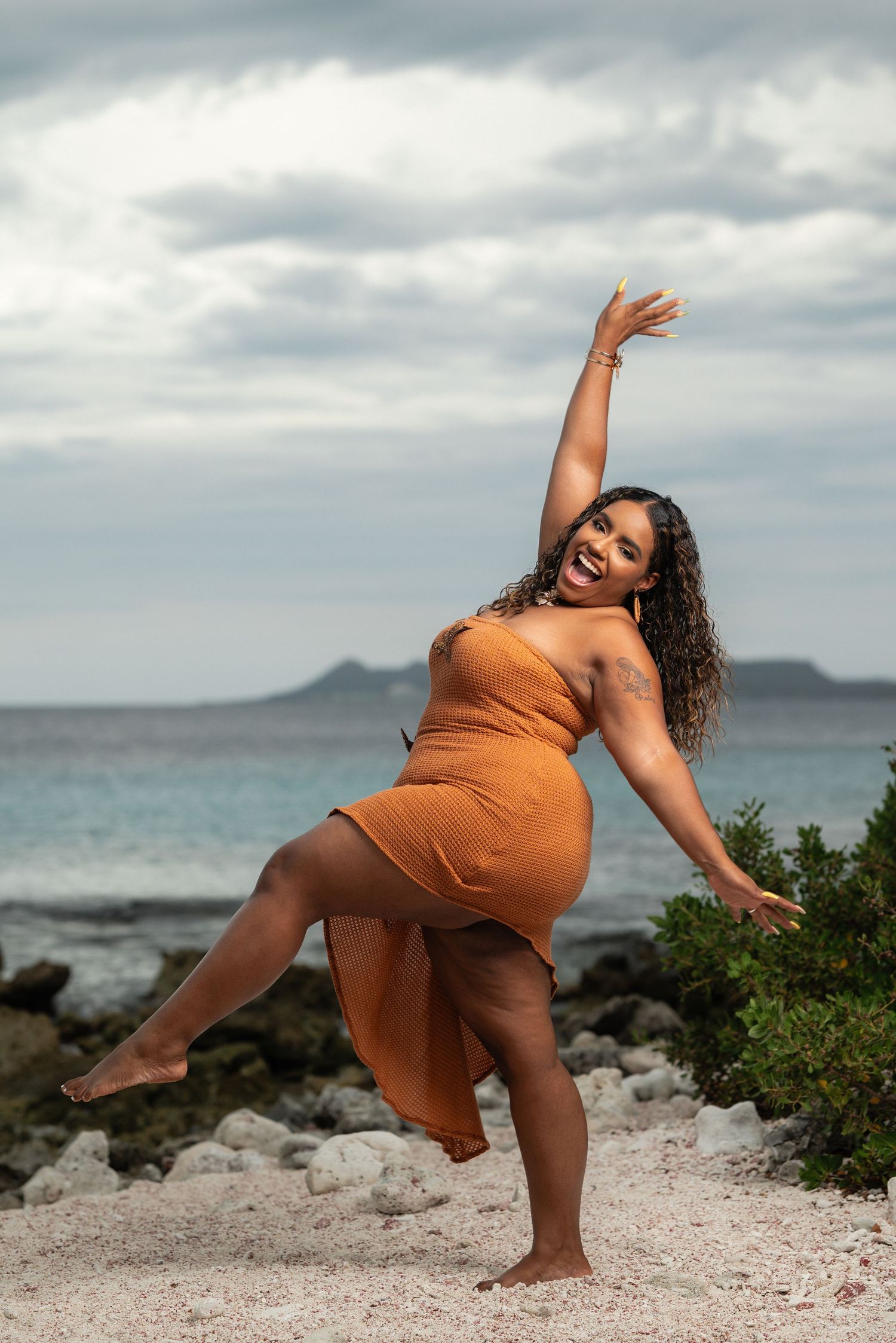 Joyful woman in orange dress dancing on beach with ocean and cloudy sky behind her.