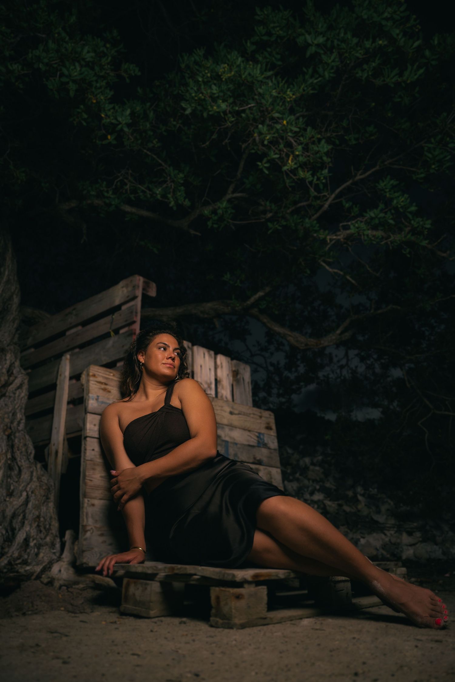 Woman in black dress sits pensively beside rustic wooden structure under trees at night.