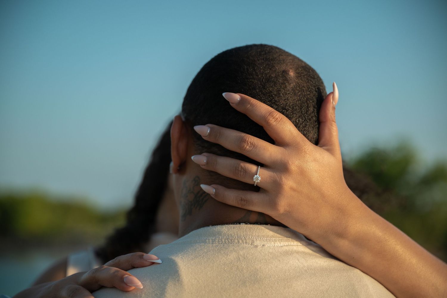 Close-up of woman with engagement ring embracing partner outdoors against blue sky.