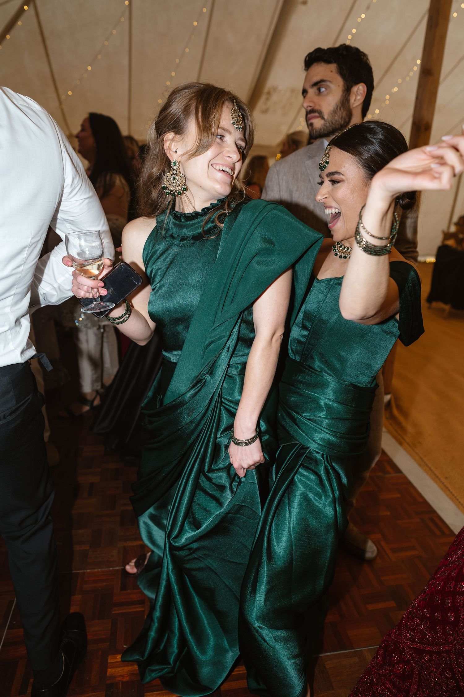Two bridesmaids in emerald green satin sarees dancing joyfully at wedding reception.