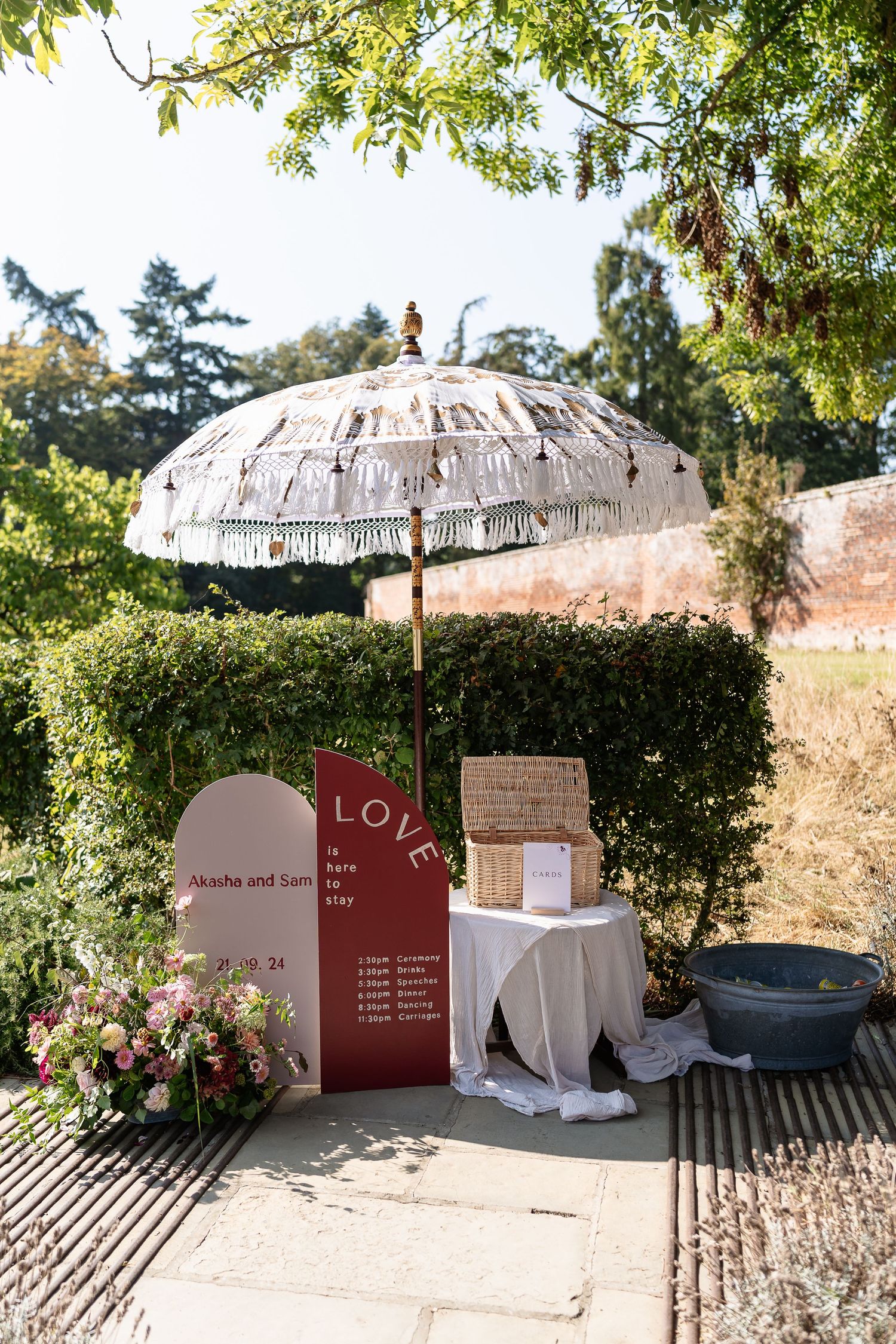 Outdoor wedding welcome sign for Akasha and Sam beneath a fringed boho umbrella in a garden.