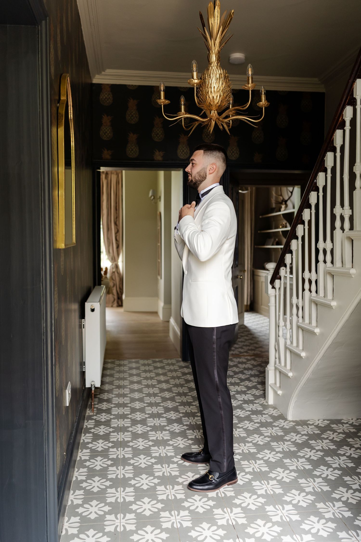 Elegant groom in white tuxedo jacket adjusting bow tie in ornate hallway.