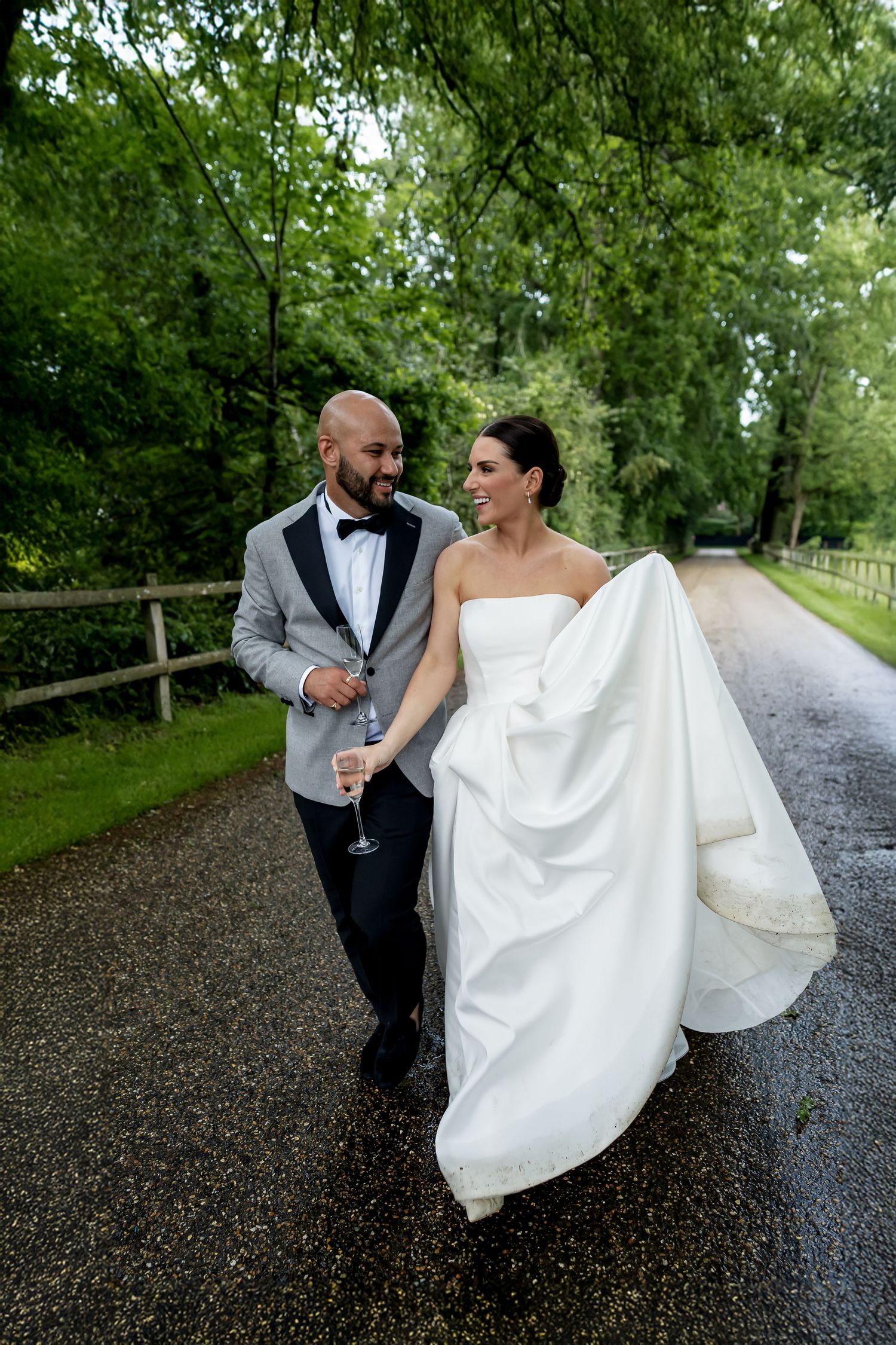 Bride and groom walk hand in hand down a tree-lined path vogue-style with groom holding champagne, bride lifting her white gown in Norfolk