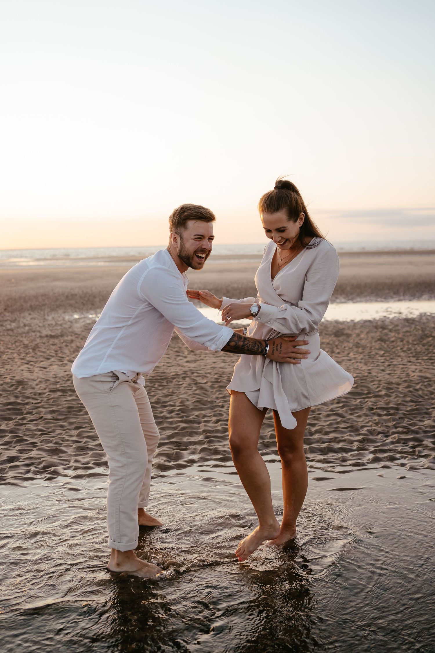 Playful couple splashing in shallow beach water at sunset, man laughing while reaching toward woman.