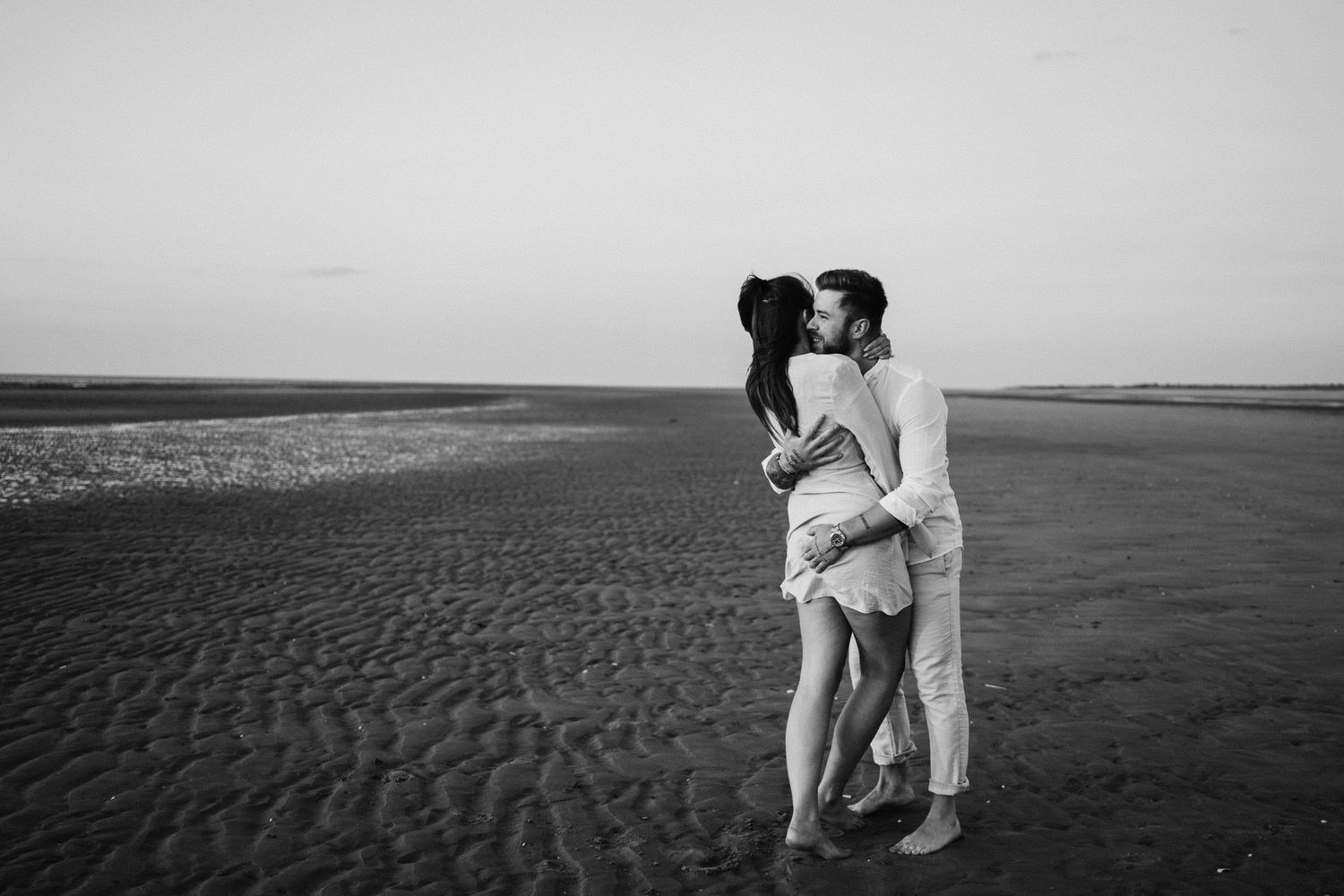 Black and white photo of a couple sharing a passionate kiss on a vast open sandy beach at dusk.