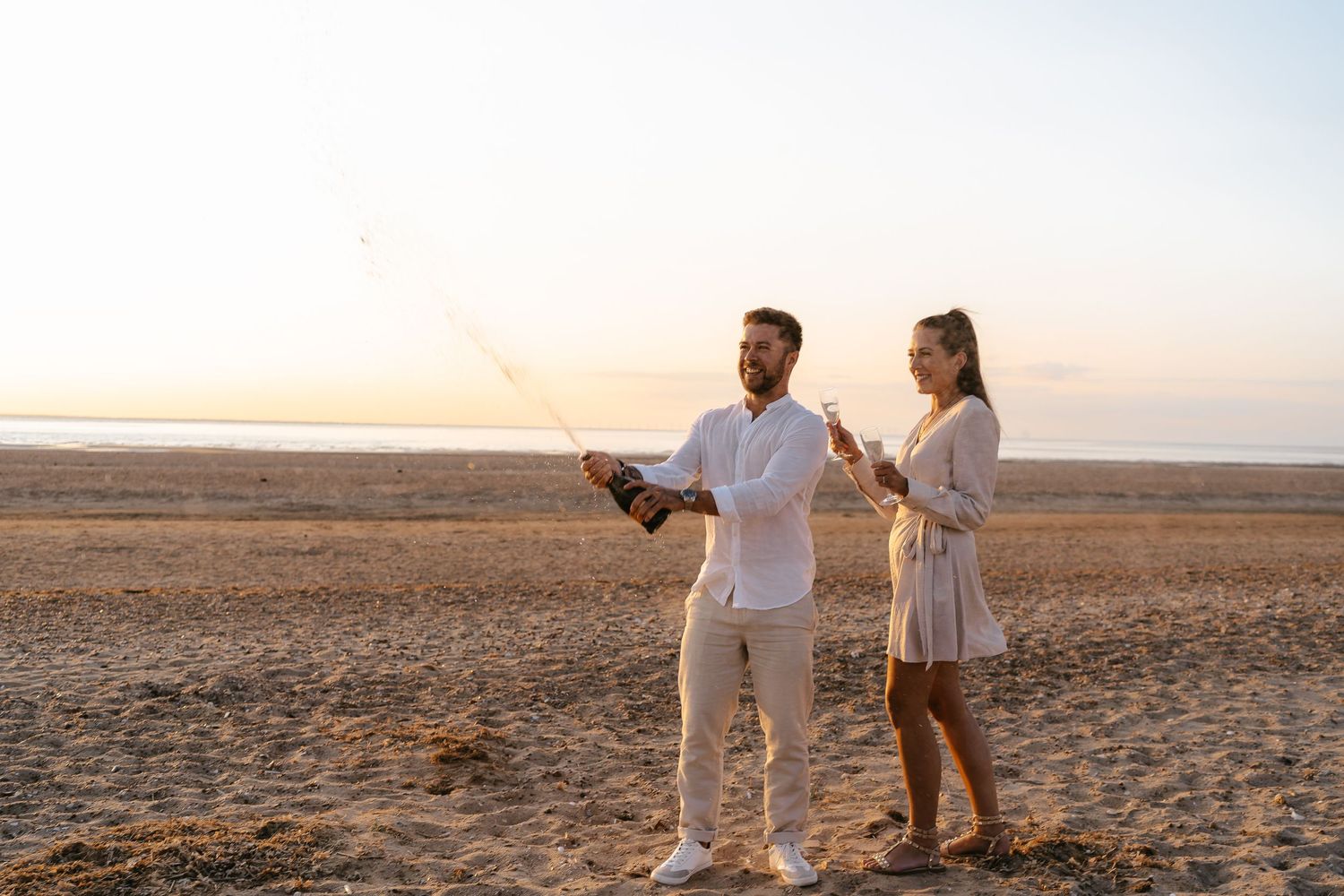 Happy couple on a beach at golden hour, man popping a bottle of champagne while woman holds a glass.