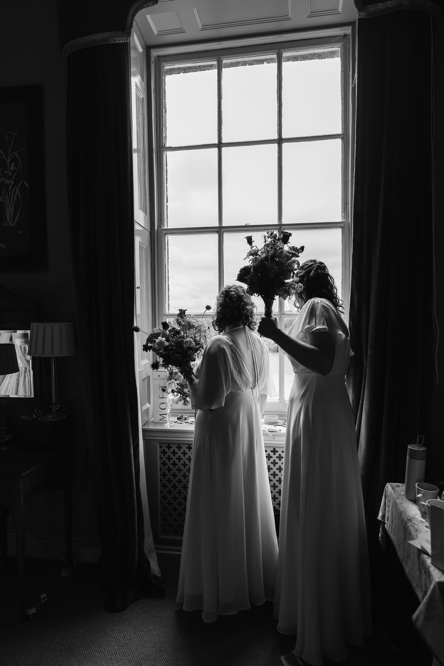 Black and white photo of two women in white dresses holding bouquets silhouetted against large window.