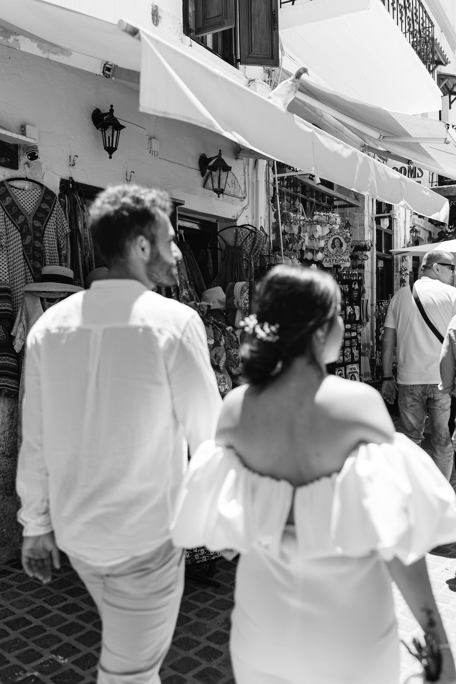 Black and white photo of newlywed couple strolling hand in hand through busy Mediterranean market street.