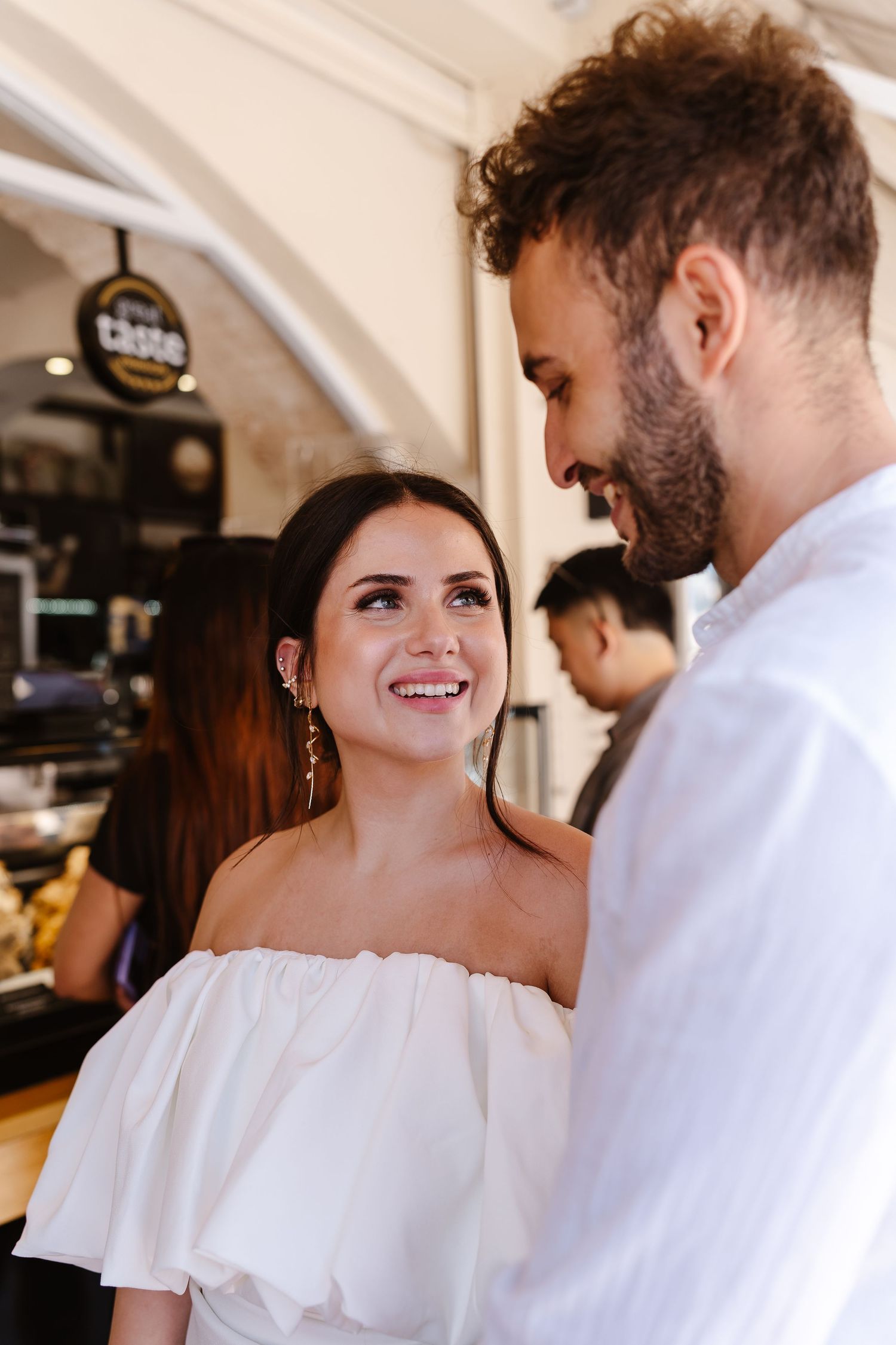 Smiling bride in white off-shoulder dress gazes lovingly at bearded groom inside a cafe.