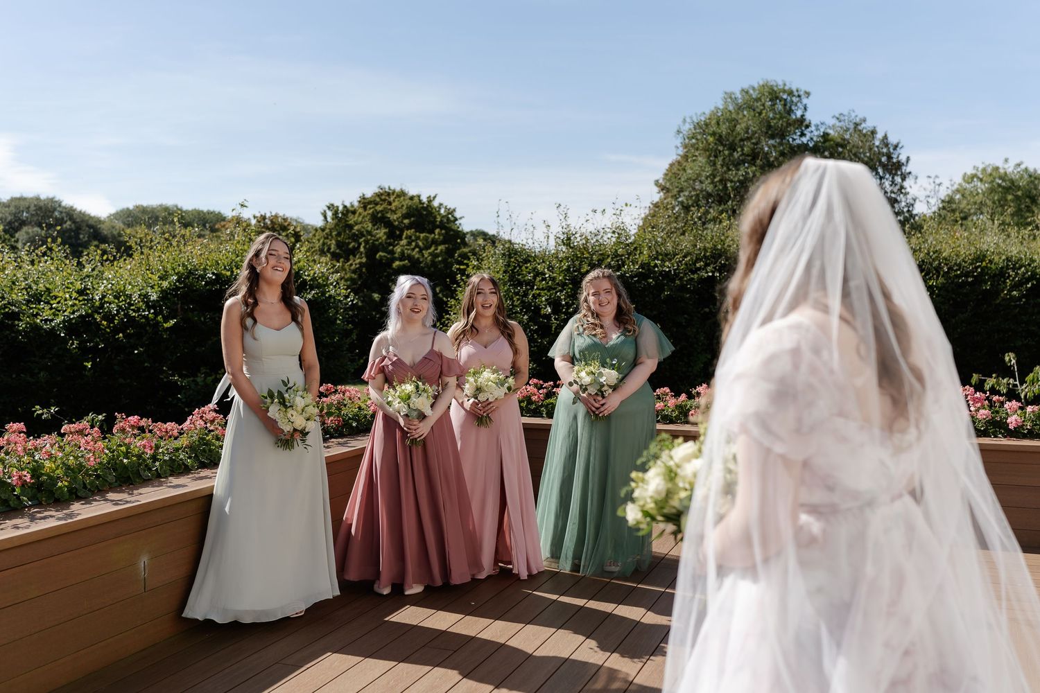 Bride in white veil reveals herself to smiling bridesmaids in pink and green dresses holding bouquets in a garden.