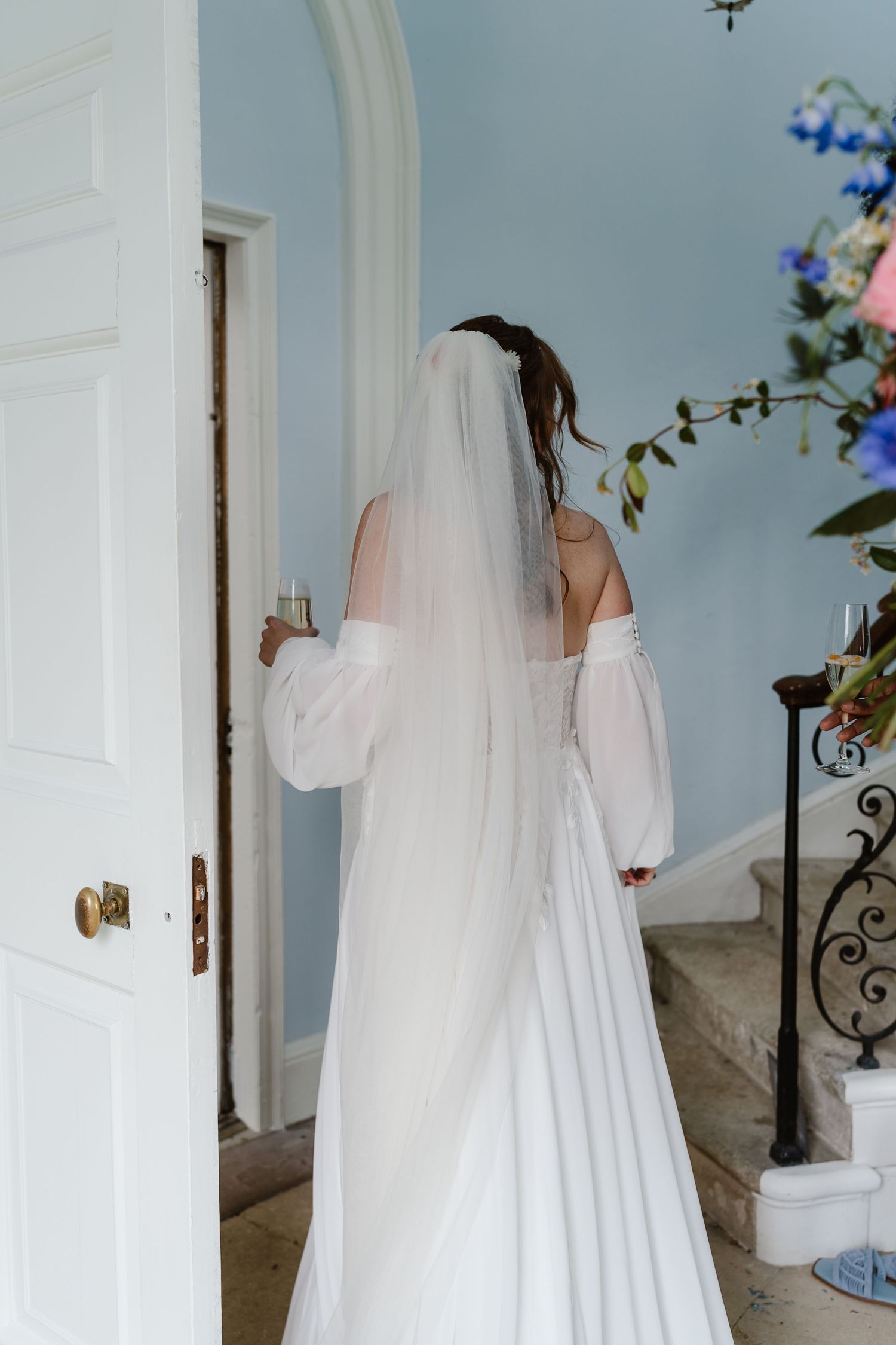 Bride in off-shoulder white gown and veil viewed from behind near an elegant doorway.