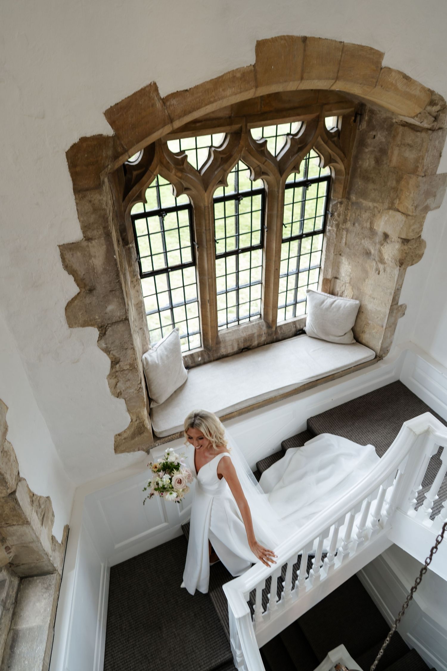 Bride in white gown holds bouquet on elegant stone staircase beneath gothic arched window in historic venue.