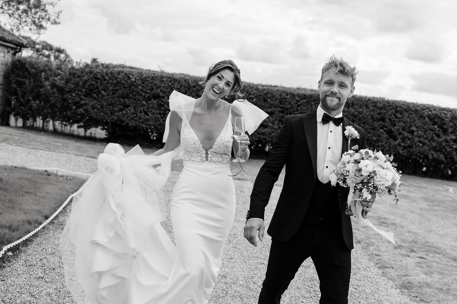Black and white photo of a laughing bride in a white gown and groom in a tuxedo walking together on their Norfolk wedding day