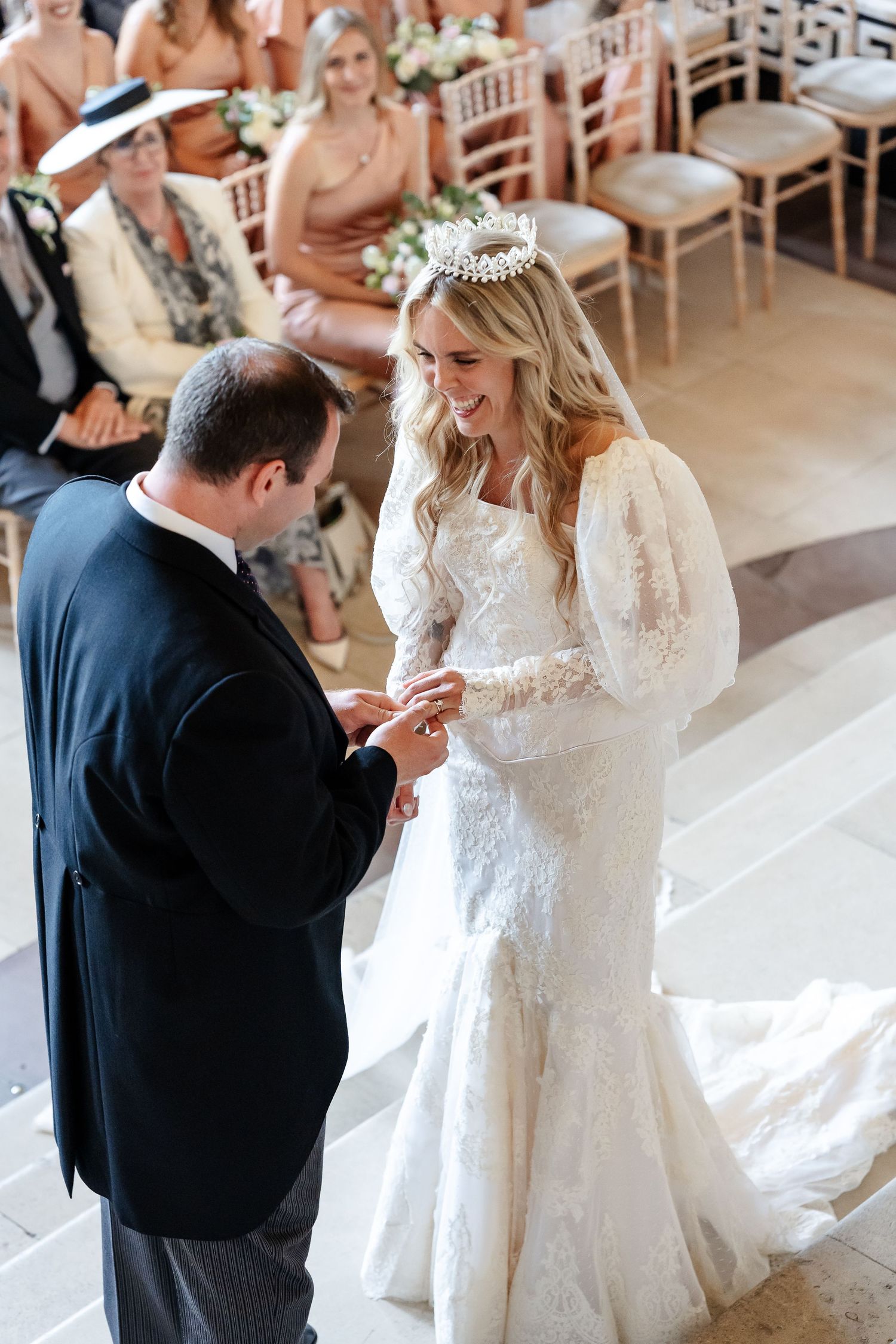 Groom placing wedding ring on bride's finger during ceremony, bride wearing lace gown and floral crown, guests seated behind.