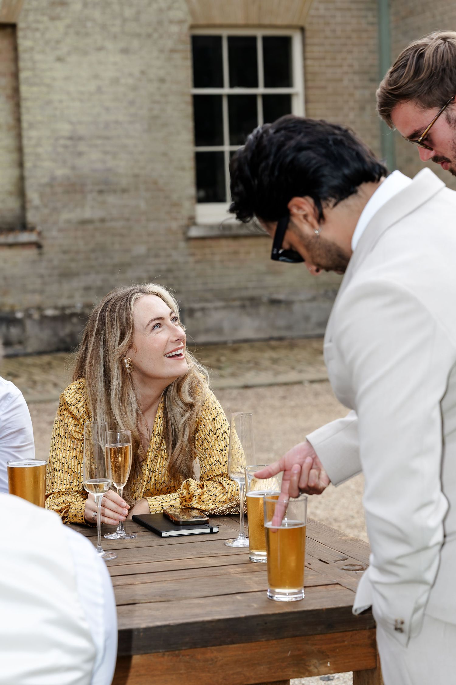 A smiling woman in a yellow floral dress enjoys champagne at an outdoor wedding reception table.