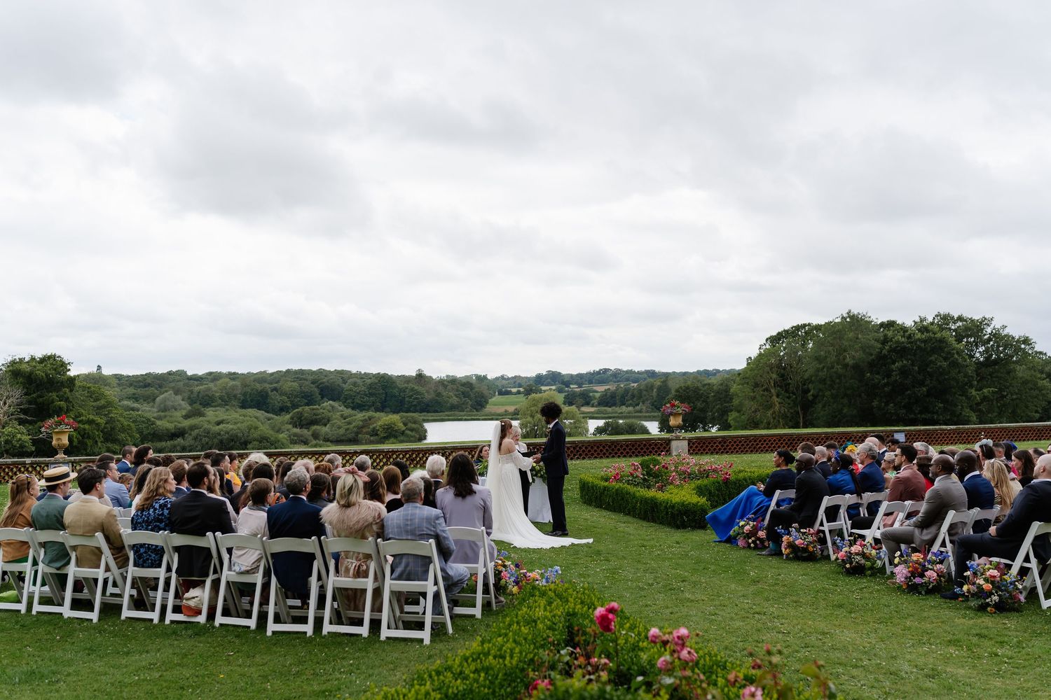 Elegant outdoor wedding ceremony with guests seated on white chairs amid lush greenery and a serene lake backdrop.