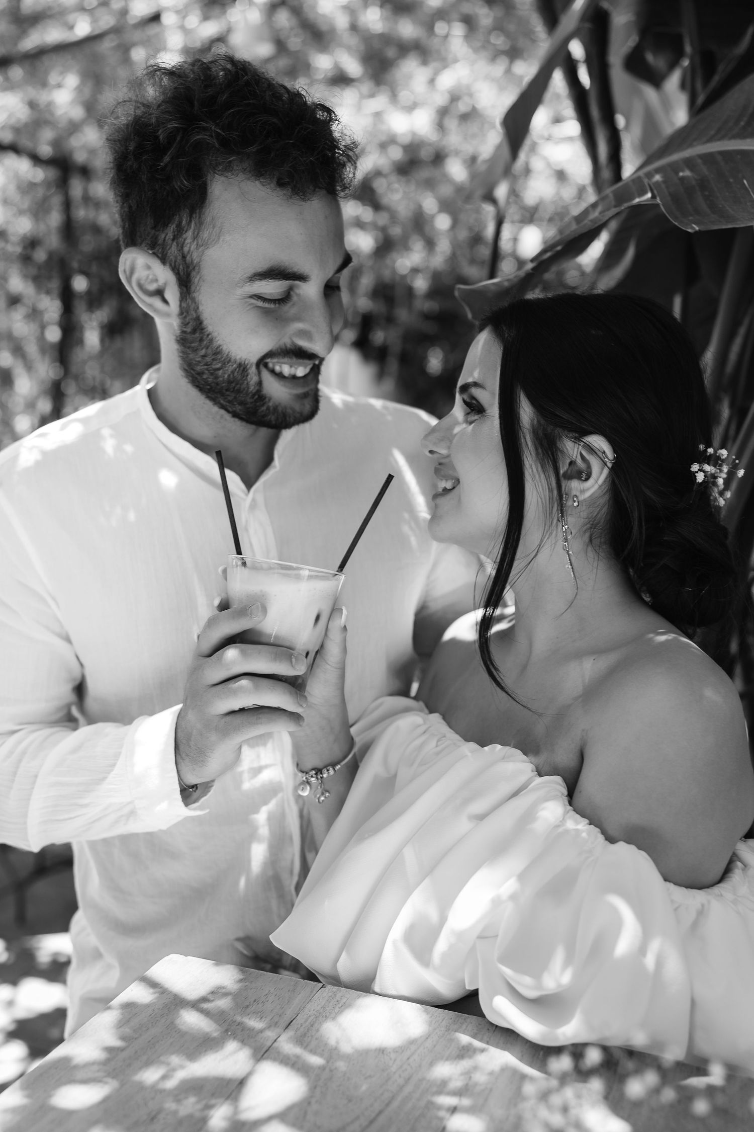 Black and white photo of a smiling couple sharing a drink at an outdoor cafe surrounded by lush trees.