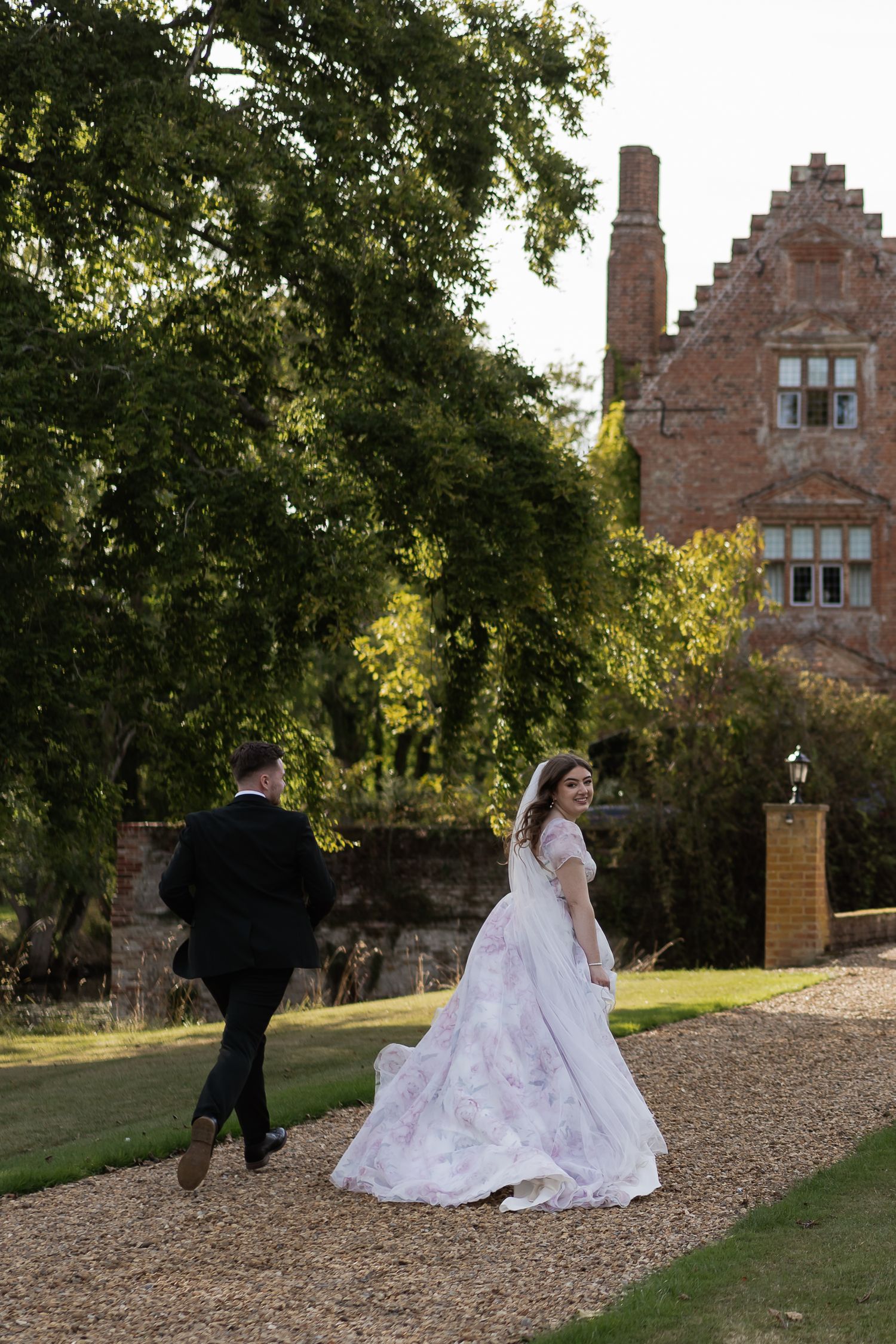Bride glances back smiling as couple strolls along garden path beside historic red brick manor.