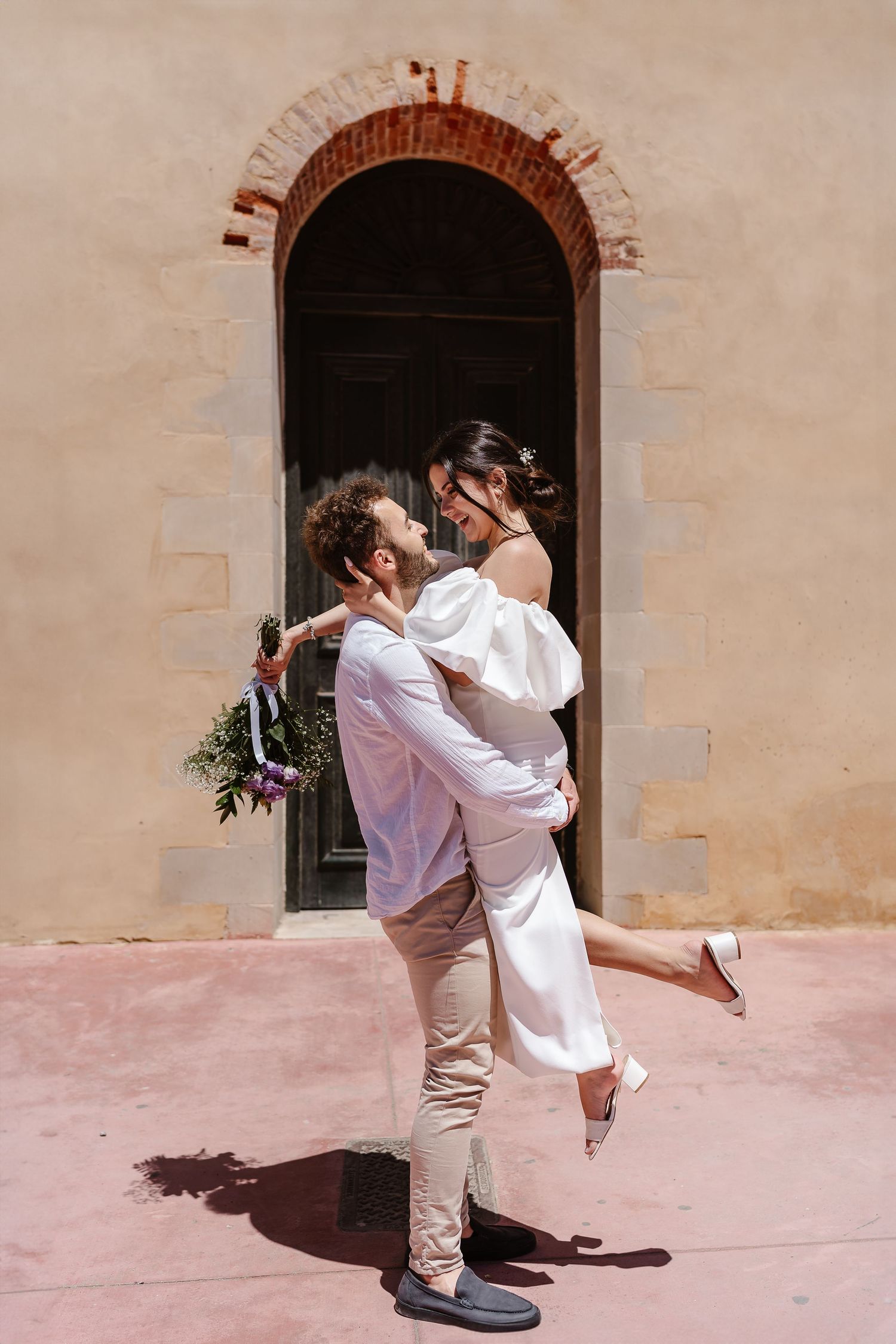 Groom lifts smiling bride in white off-shoulder dress outside a rustic arched doorway in a sun-drenched Mediterranean setting.