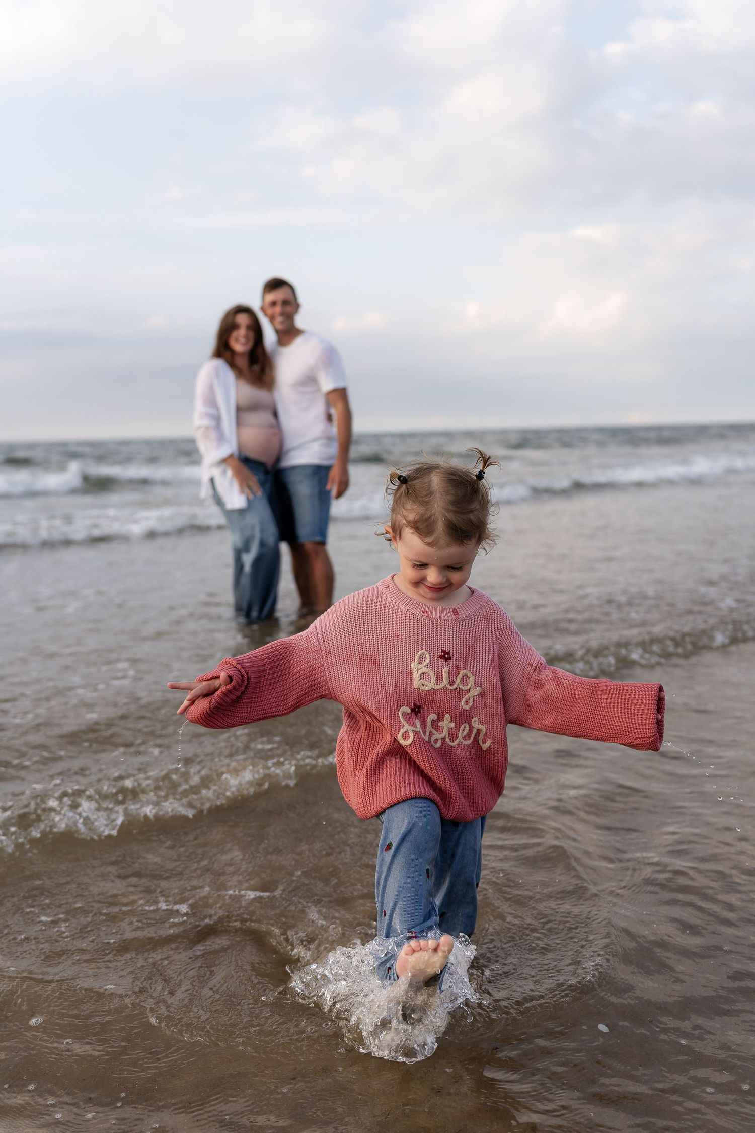 Young girl in Big Sister sweater splashes in ocean waves as expecting parents watch from behind.