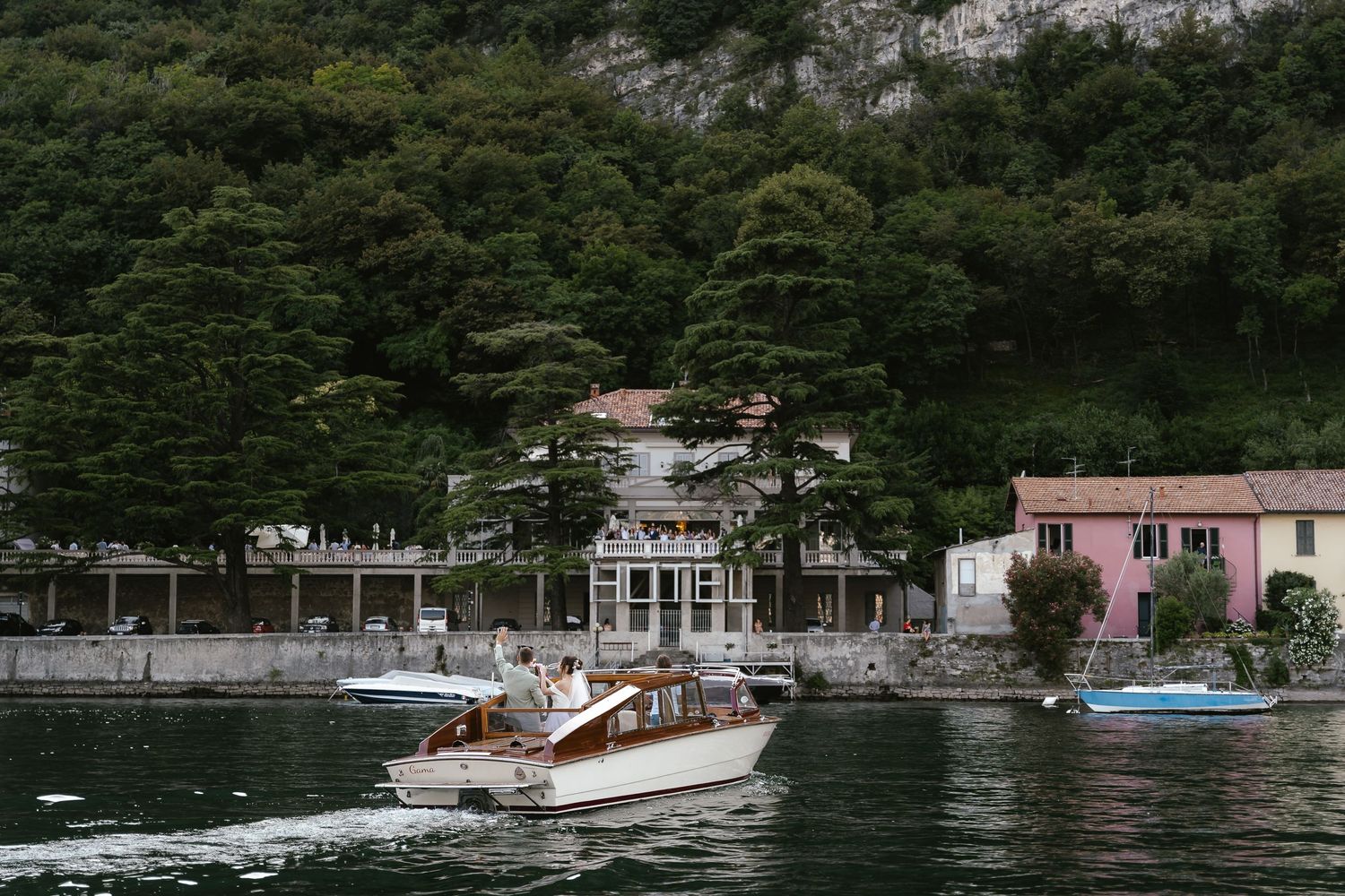 Classic wooden boat cruising past elegant lakeside villa and pink building on scenic lake.